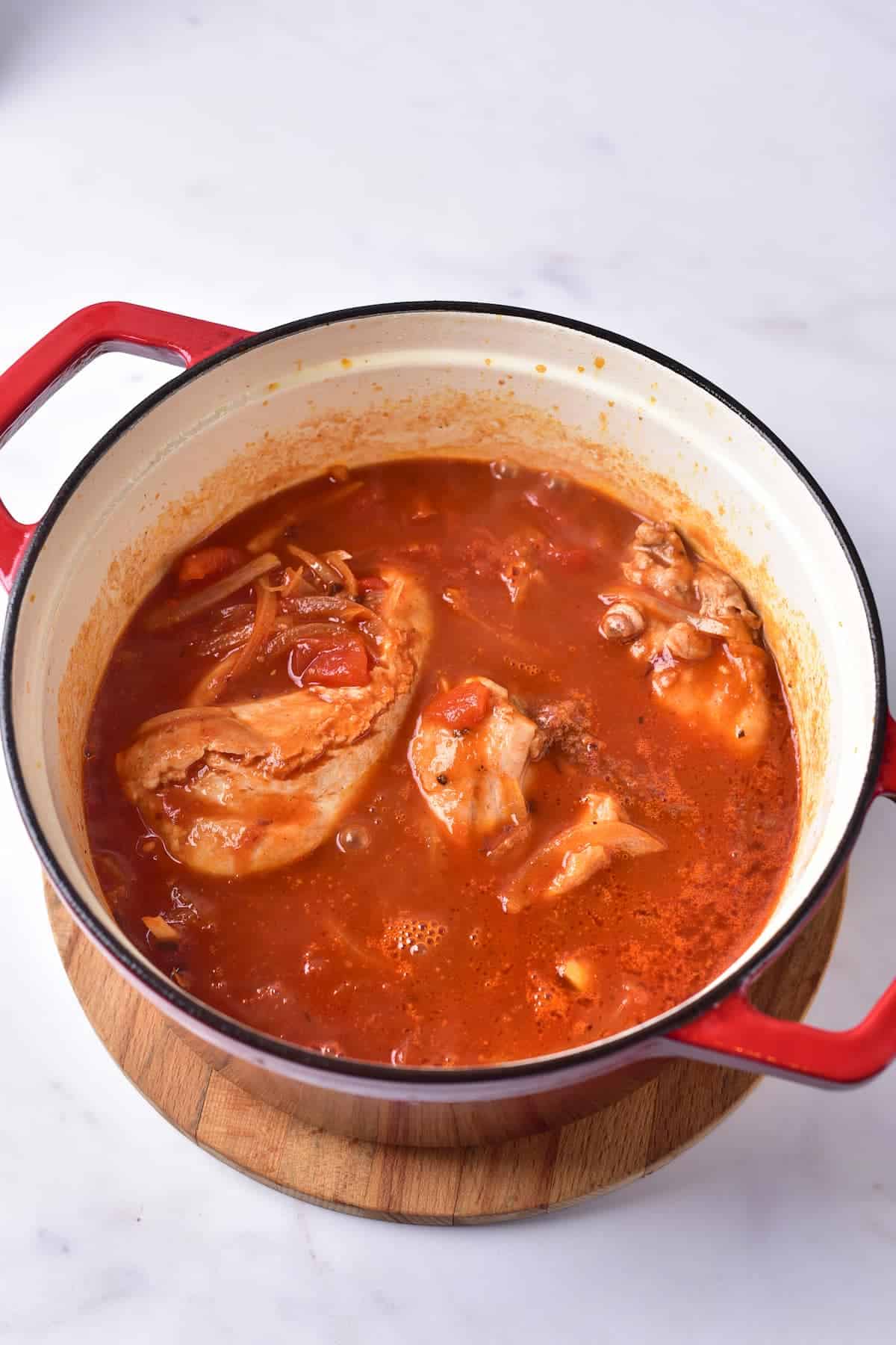 Red pot with chicken pieces simmering in tomato-based stew on a wooden trivet, photographed from above on a white surface.