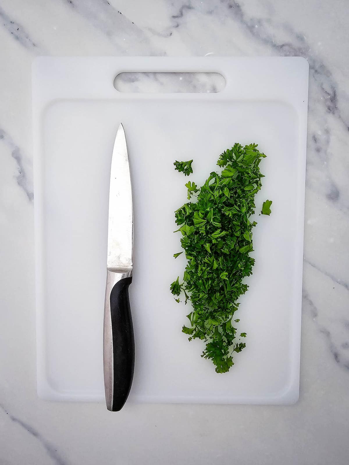 A knife and a pile of chopped fresh parsley are on a white cutting board placed on a marble countertop.