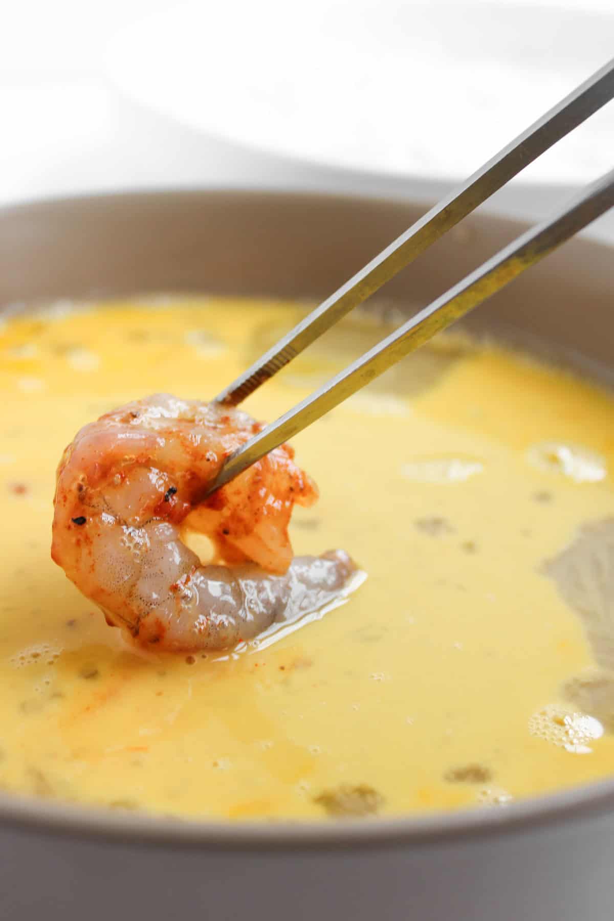 A pair of metal chopsticks holds a raw, seasoned shrimp above a bowl of beaten eggs.