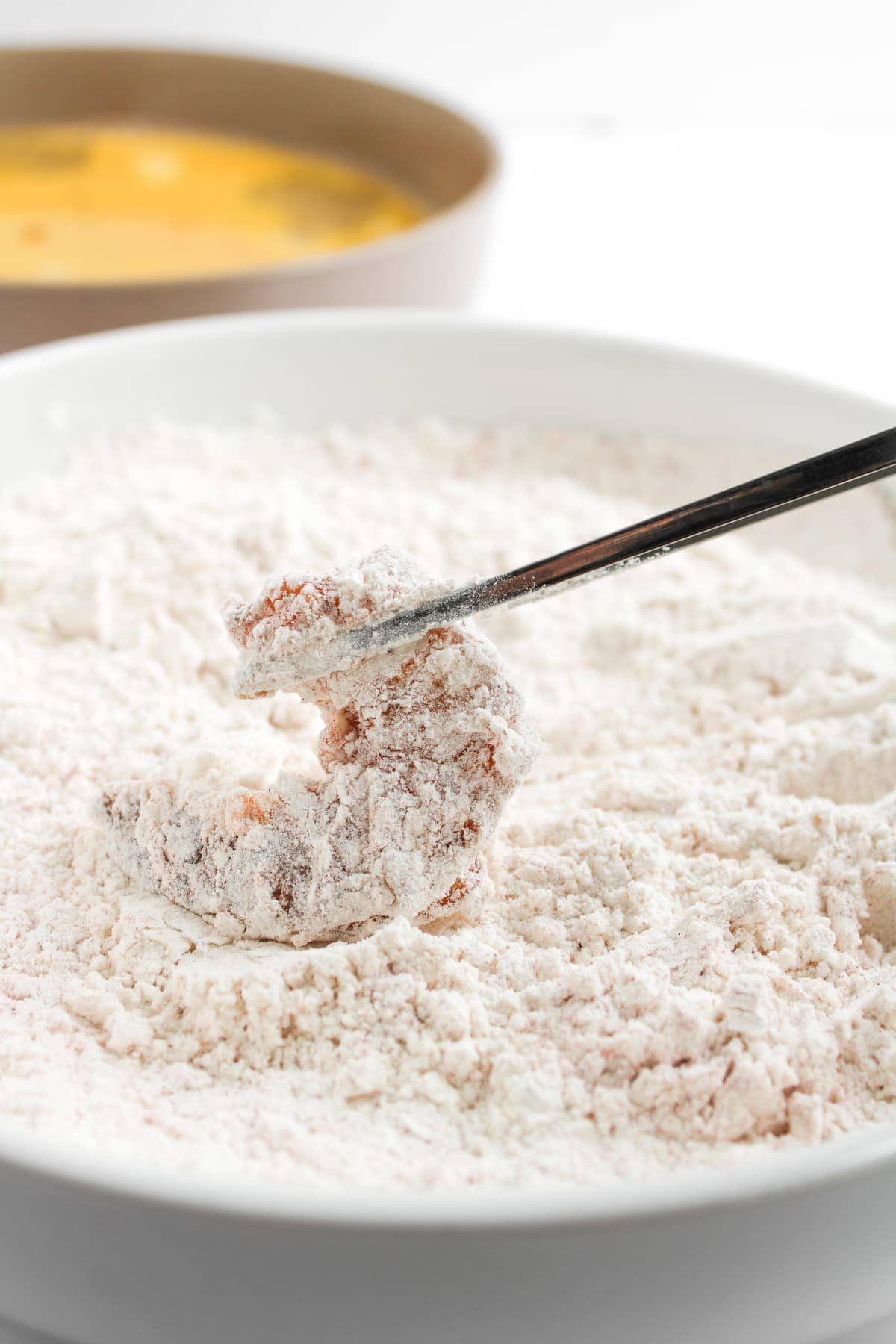 A piece of raw meat is being coated in flour with chopsticks; a bowl with a yellow liquid mixture is in the background.