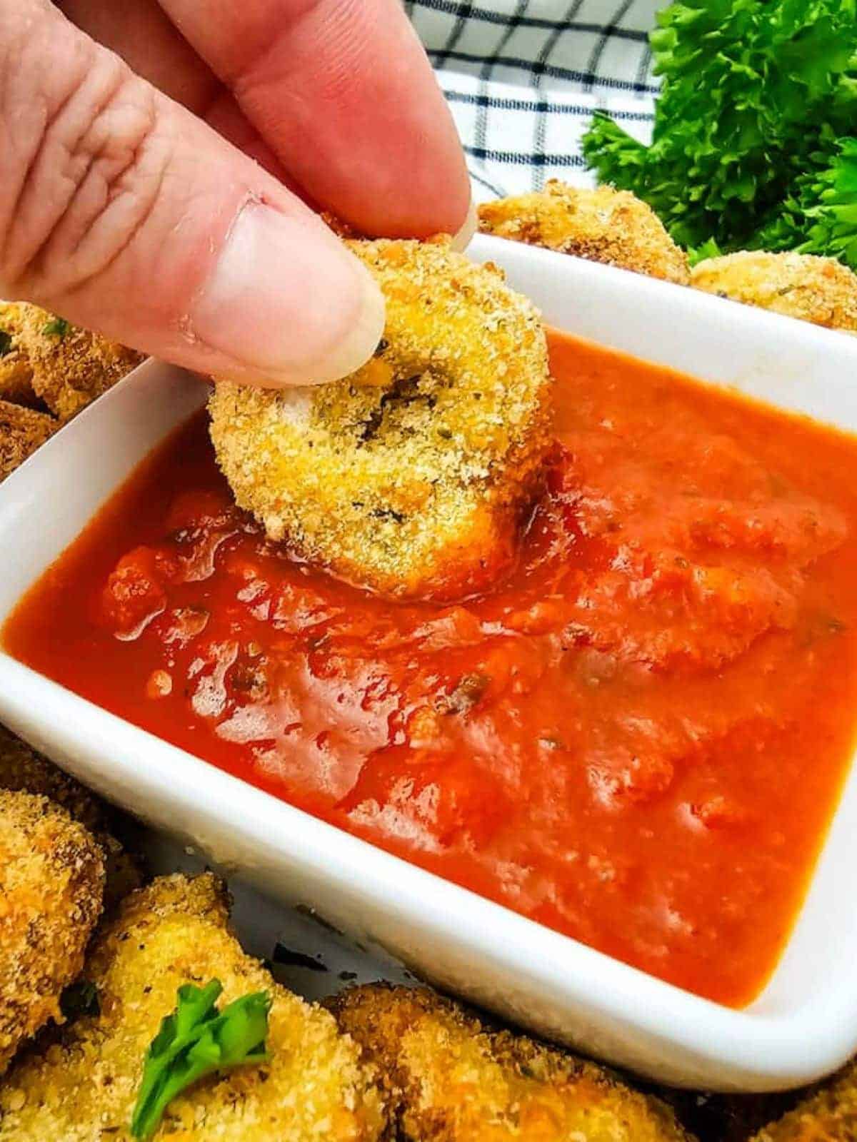 A hand dips a breaded, fried mushroom into a bowl of marinara sauce, with more fried mushrooms and parsley in the background.