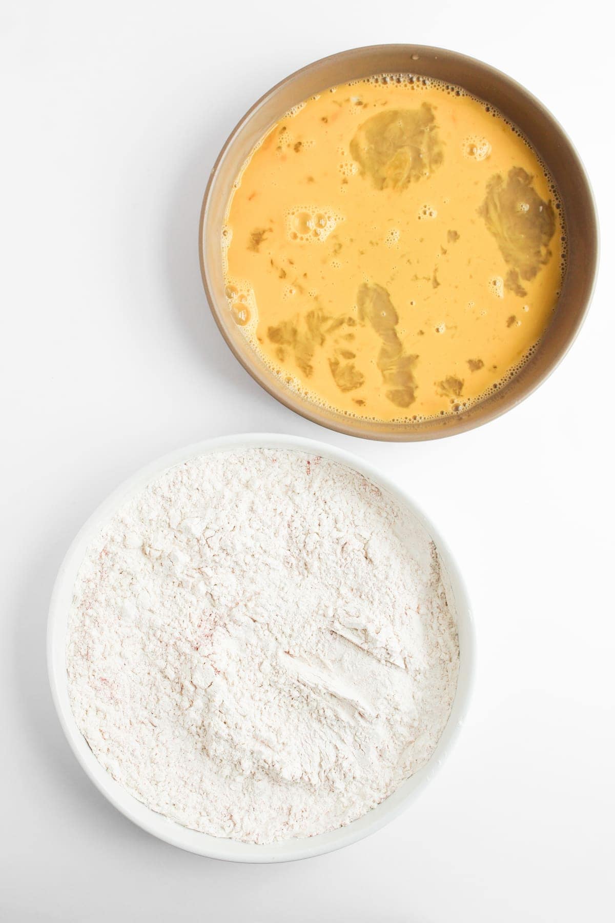 Two bowls on a white surface: one containing a beaten egg mixture, the other filled with flour.