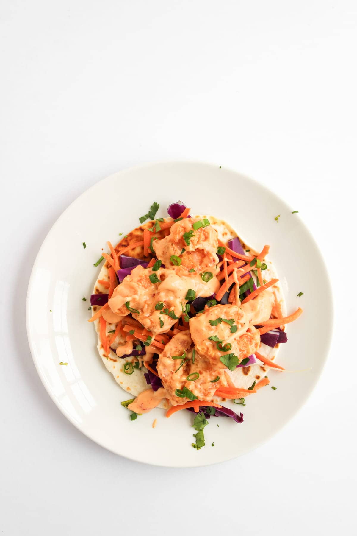 A plate with shrimp on a flatbread, topped with shredded carrots, purple cabbage, and chopped herbs, on a white background.