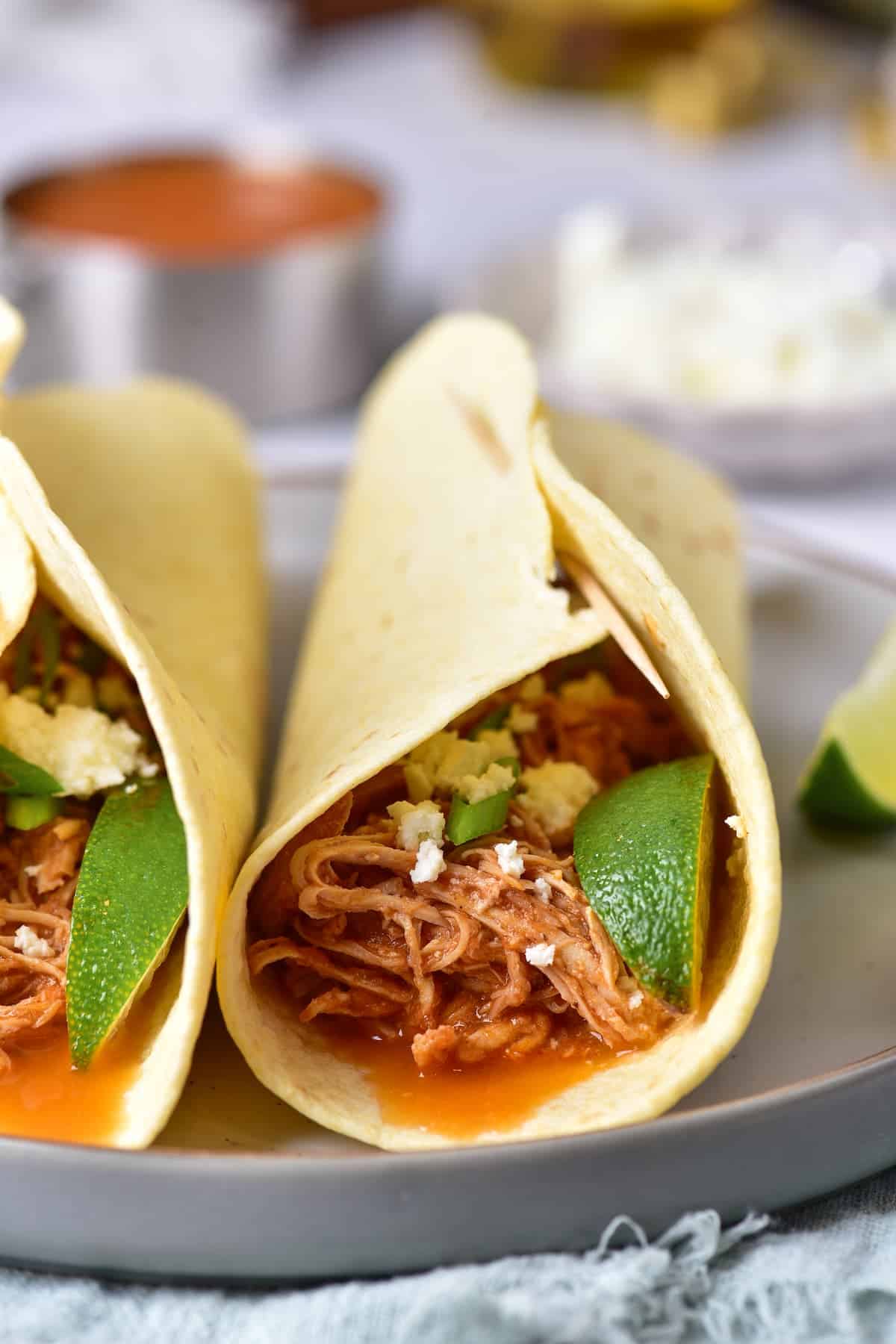 Two soft tacos filled with shredded meat, crumbled cheese, sliced jalapeños, and lime wedges on a gray plate, with sauce and cheese in the background.