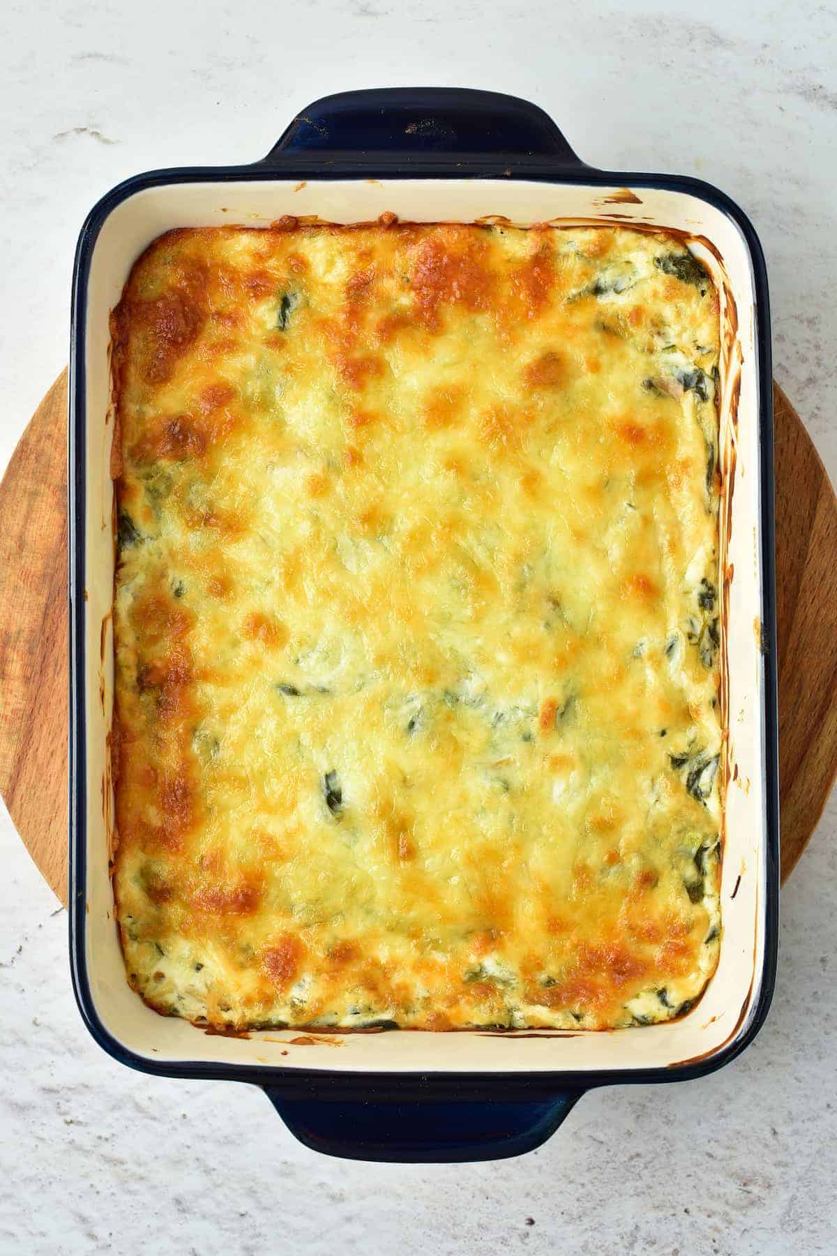 A baked casserole in a rectangular dish with a golden-brown, melted cheese topping, set on a wooden board.