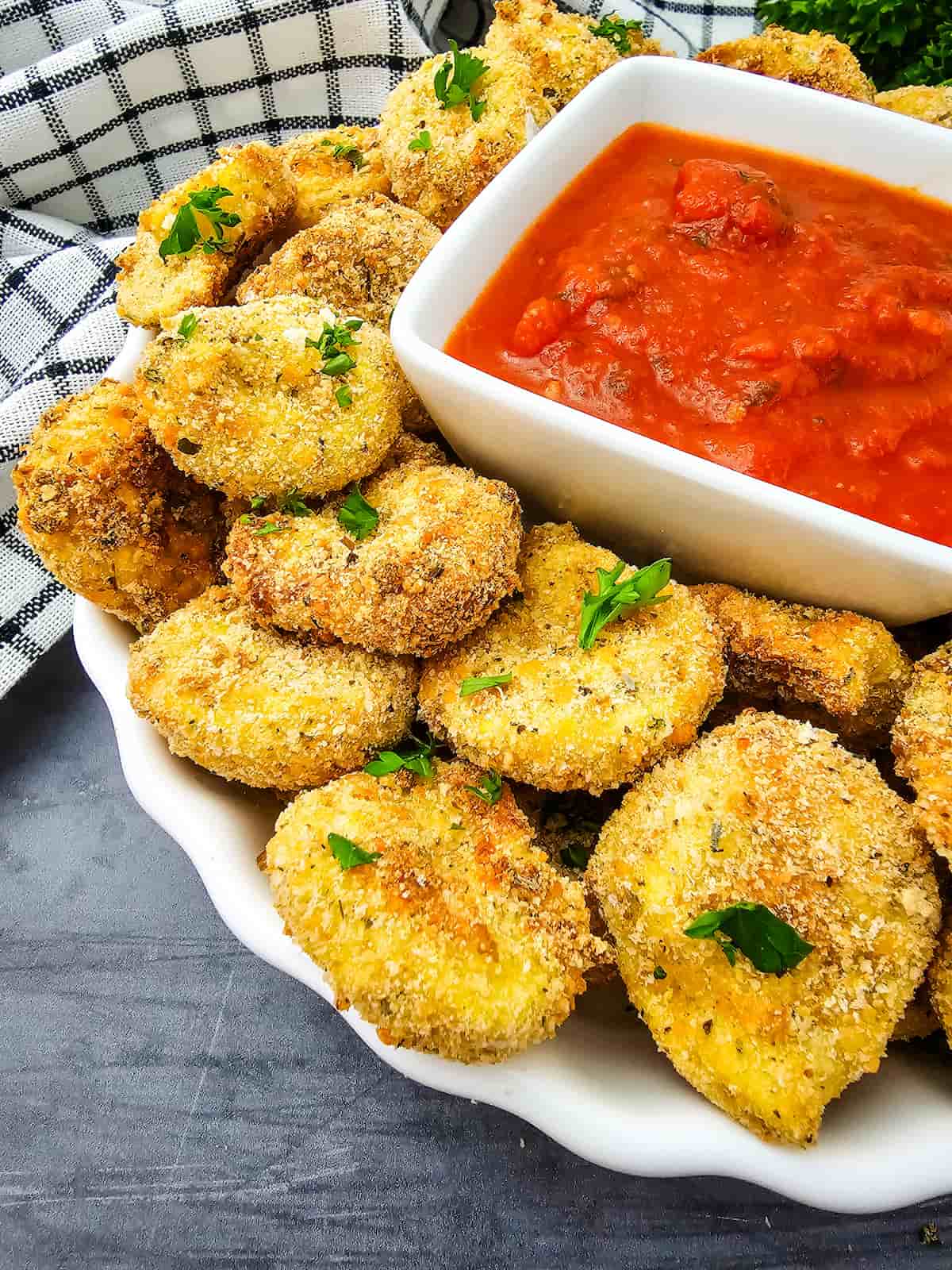 A plate of breaded, baked zucchini chips garnished with parsley, served with a bowl of chunky tomato dipping sauce.