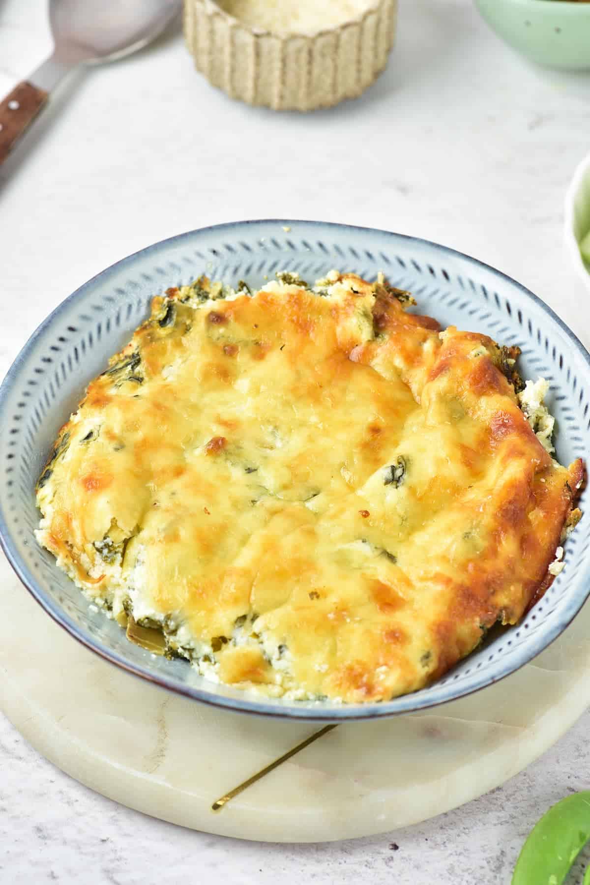 A baked spinach and cheese casserole in a round blue dish, resting on a white marble surface.