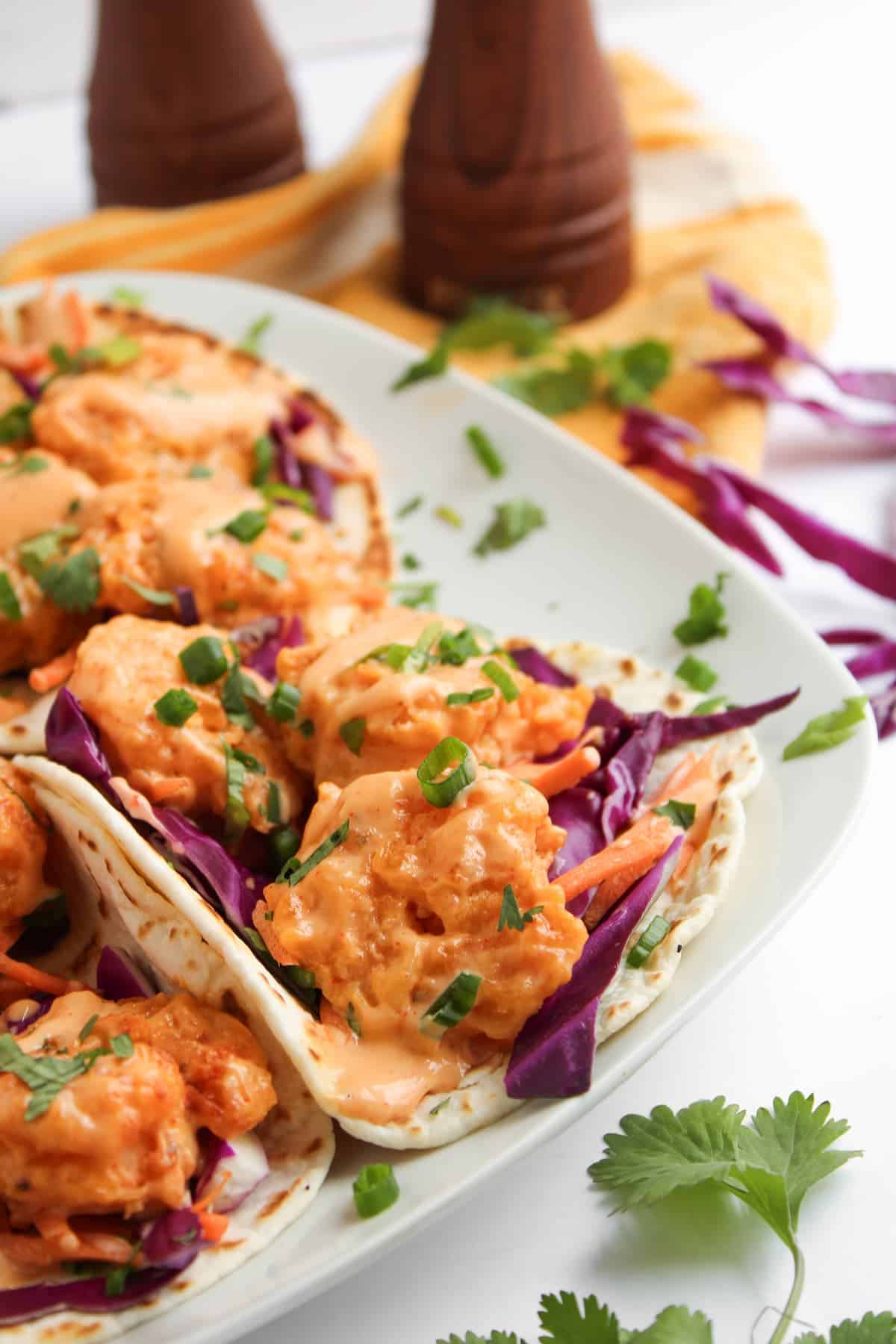 Three soft tacos filled with fried shrimp, purple cabbage, shredded carrots, and creamy orange sauce, garnished with chopped green onions on a white plate.