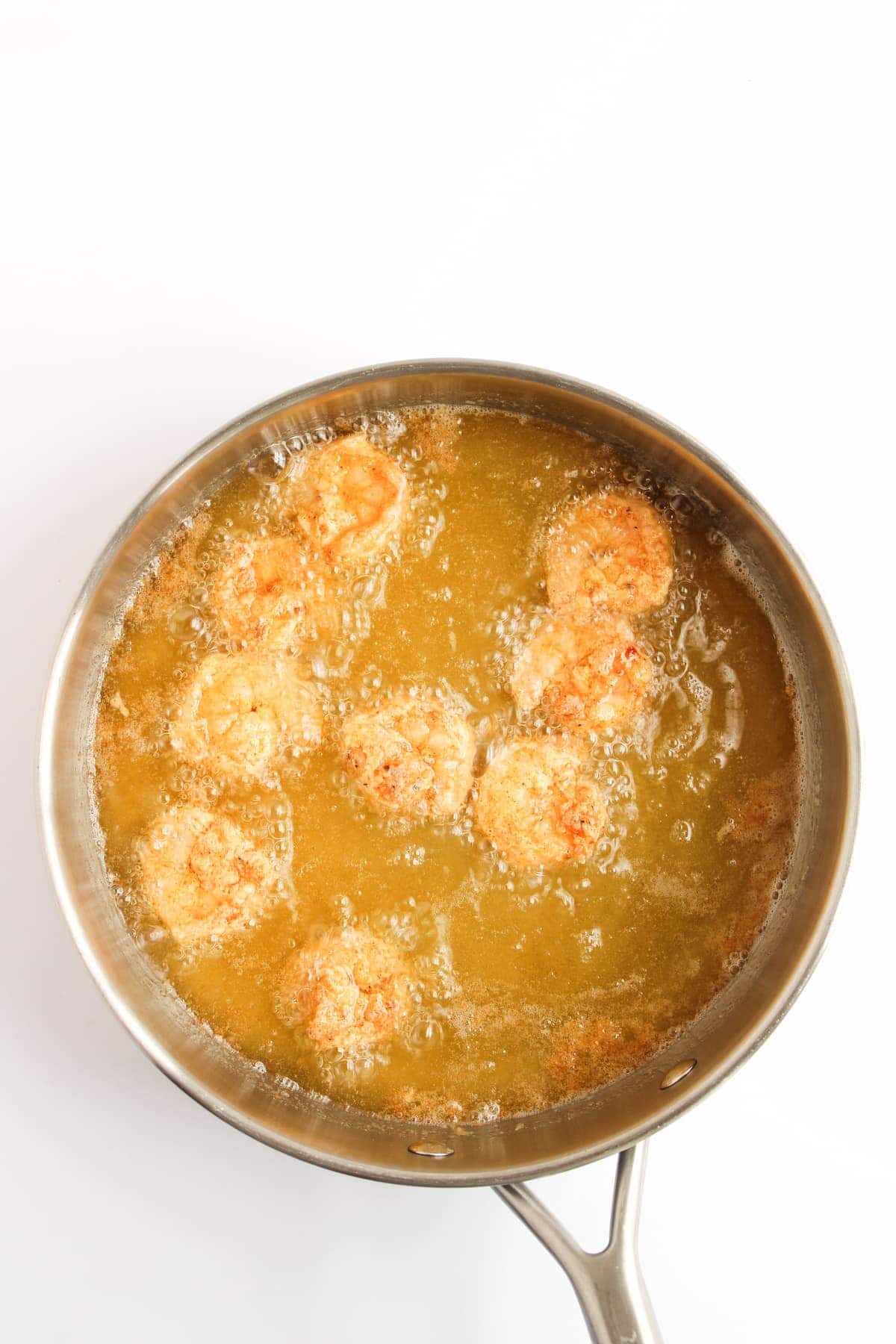 A stainless steel pan filled with oil, frying several breaded shrimp with bubbles forming around them.