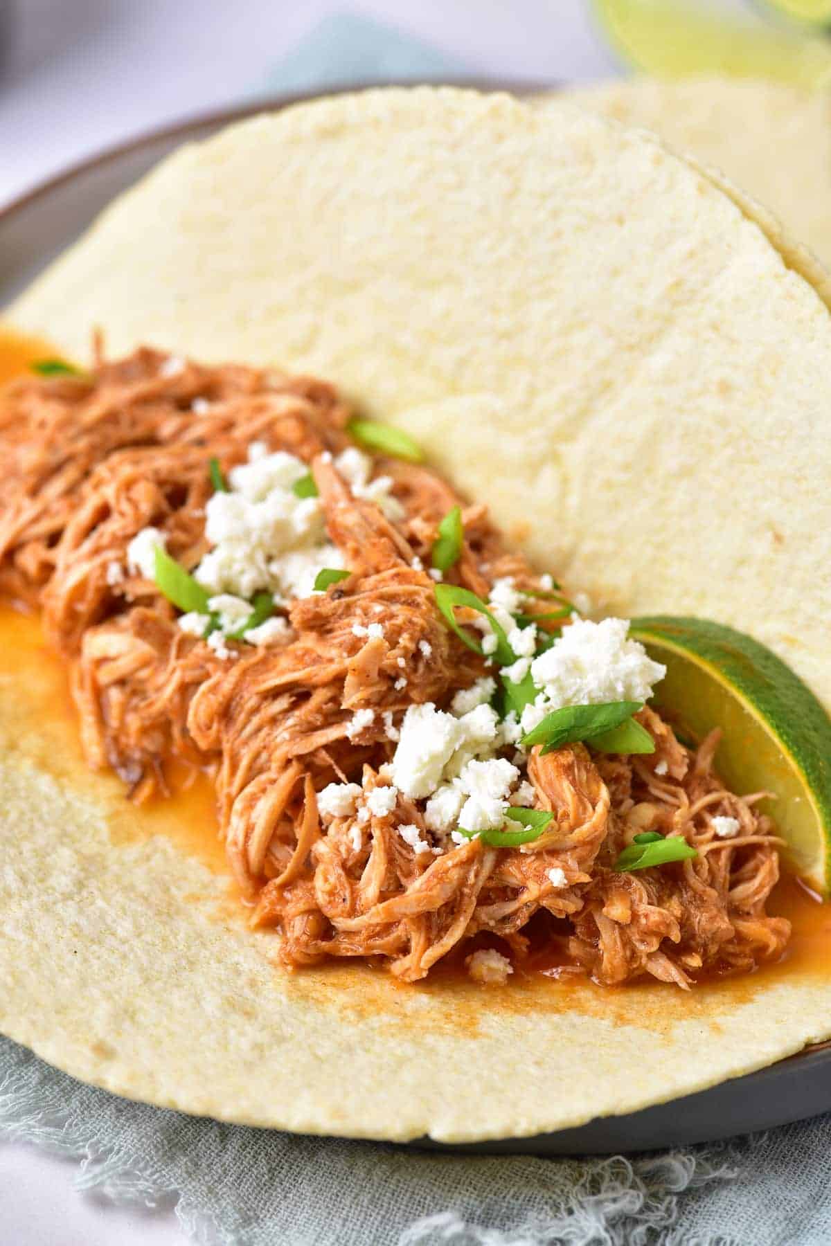 A corn tortilla filled with shredded chicken in red sauce, garnished with crumbled cheese, green onions, and a lime wedge.