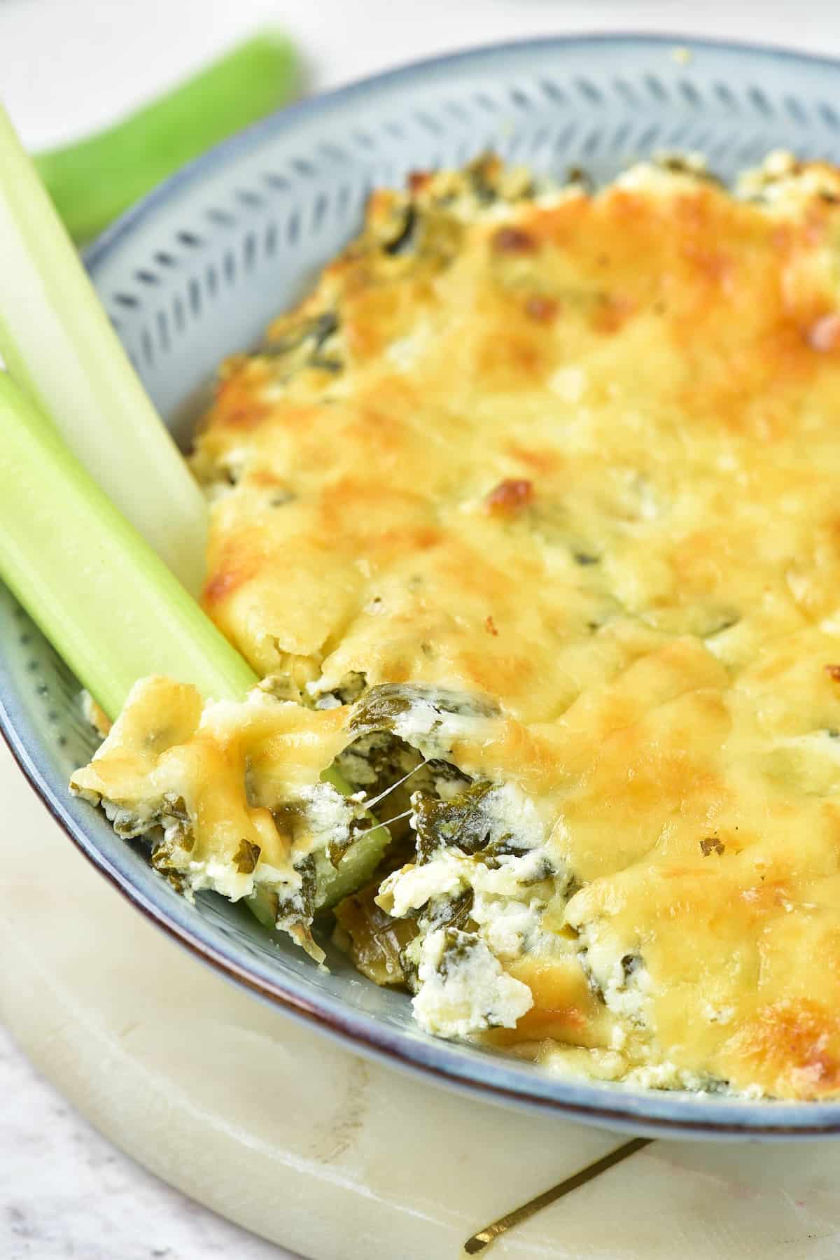 A bowl of baked spinach and cheese dip, partially served with a side of fresh celery sticks.