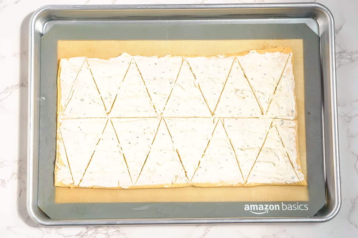 A baking sheet lined with a silicone mat holds a large rectangle of dough spread with a creamy herb mixture, cut into triangles.