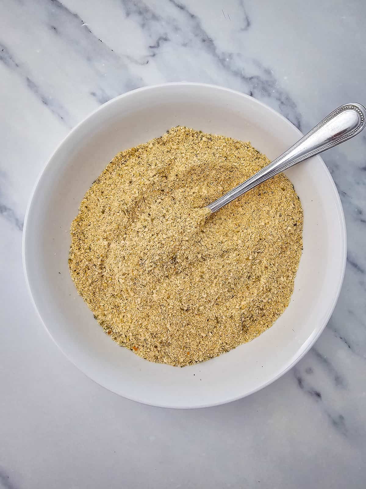 A white bowl filled with a beige, finely ground seasoning blend, with a metal spoon resting inside, on a marble surface.