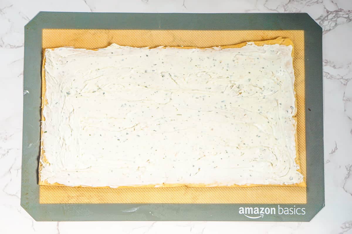 A rectangular baking mat topped with a sheet of pastry evenly spread with a creamy white herb mixture, on a marble surface.