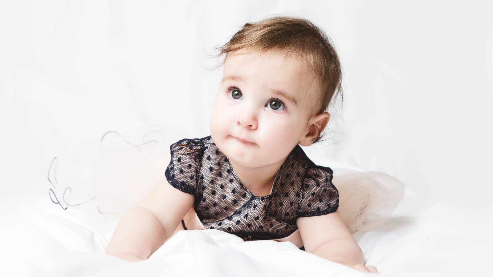 A baby wearing a navy blue polka dot dress lies on a white blanket, looking slightly upward with a neutral expression.