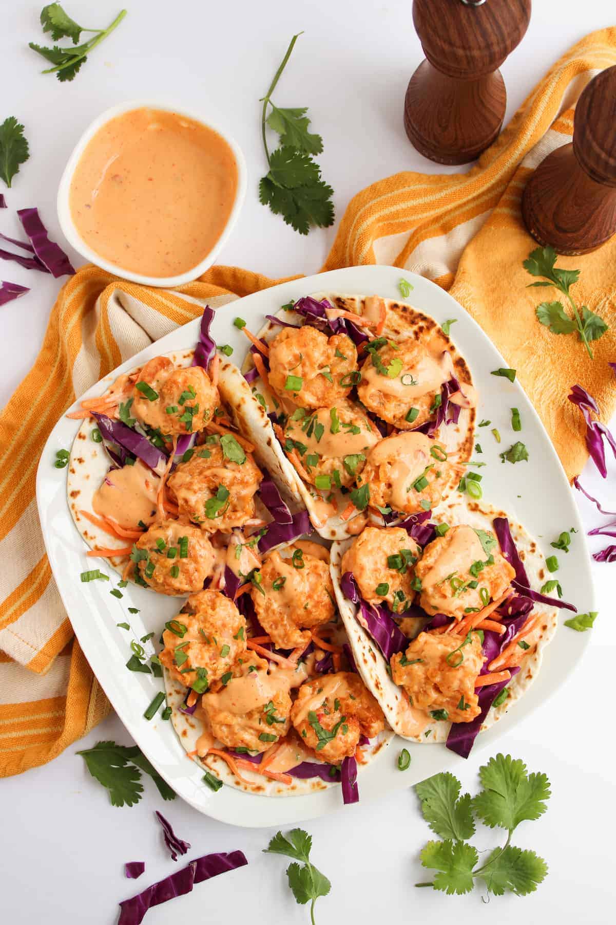 A plate of shrimp tacos topped with sauce, chopped herbs, and sliced cabbage, served with extra sauce on the side and garnished with cilantro.
