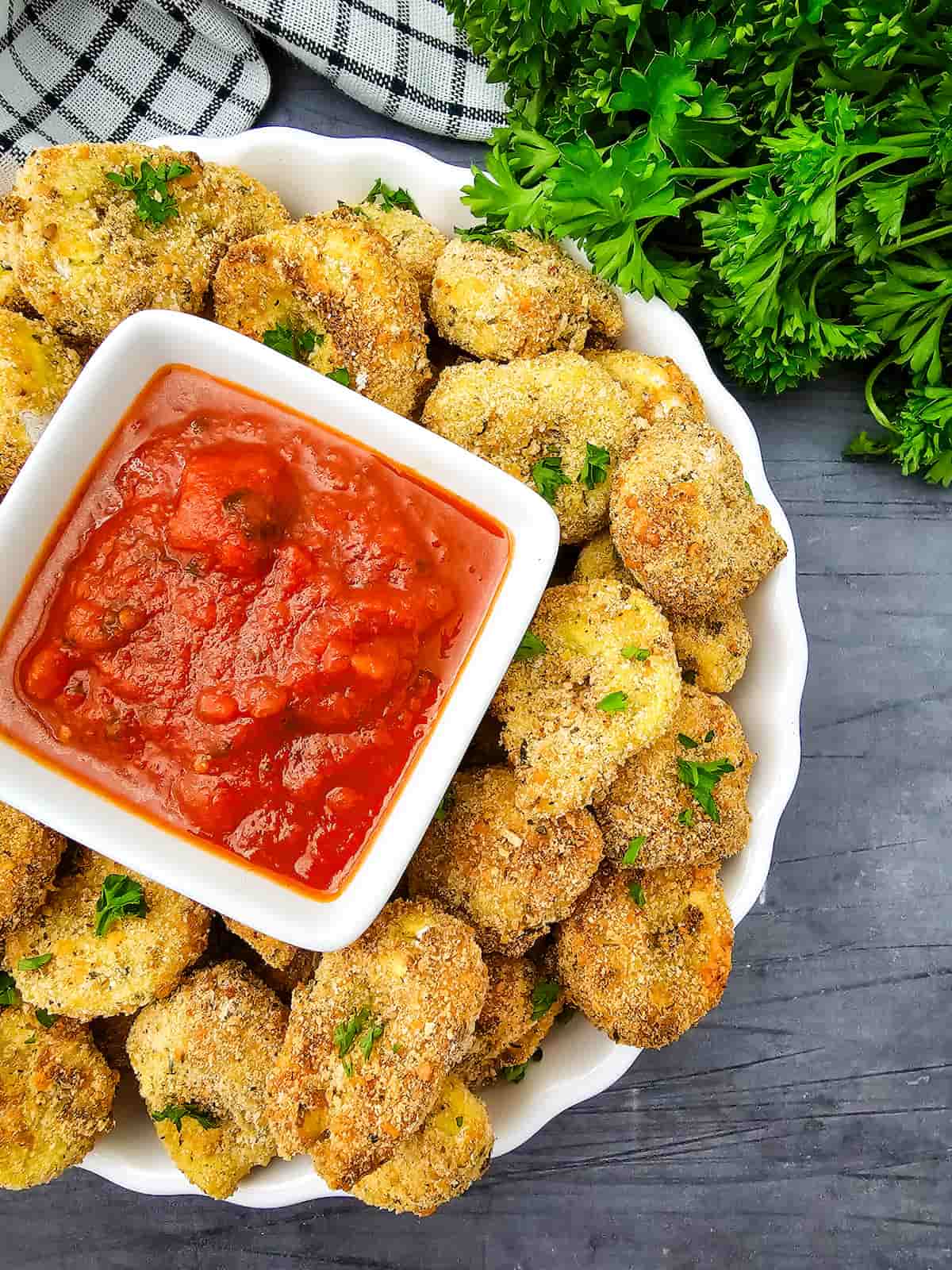 A white plate of breaded, baked nuggets garnished with parsley, served with a bowl of tomato dipping sauce on a gray table next to fresh herbs.