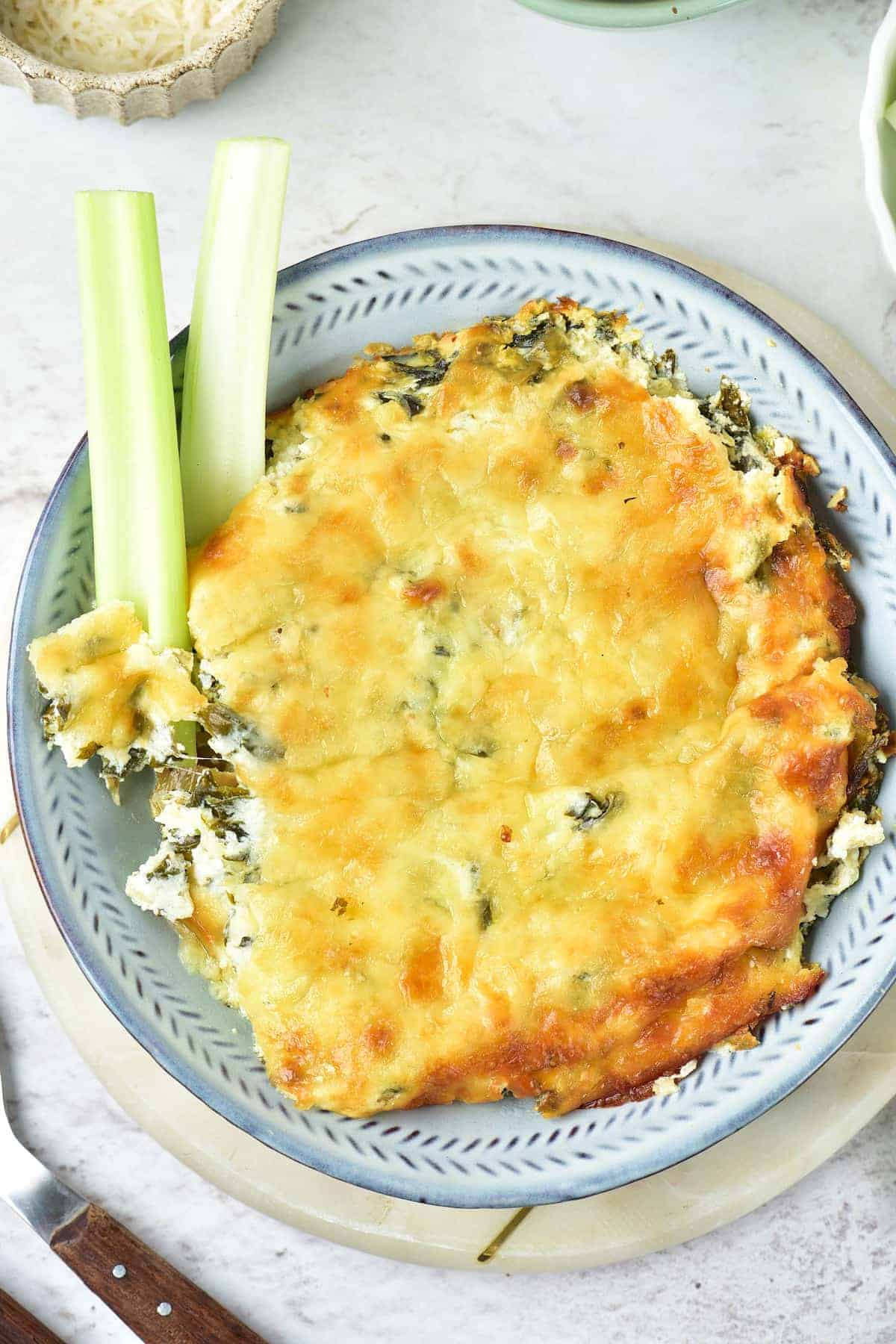 A plate of baked cheesy spinach dip with two celery sticks placed on the side, partially dipped in the dish.