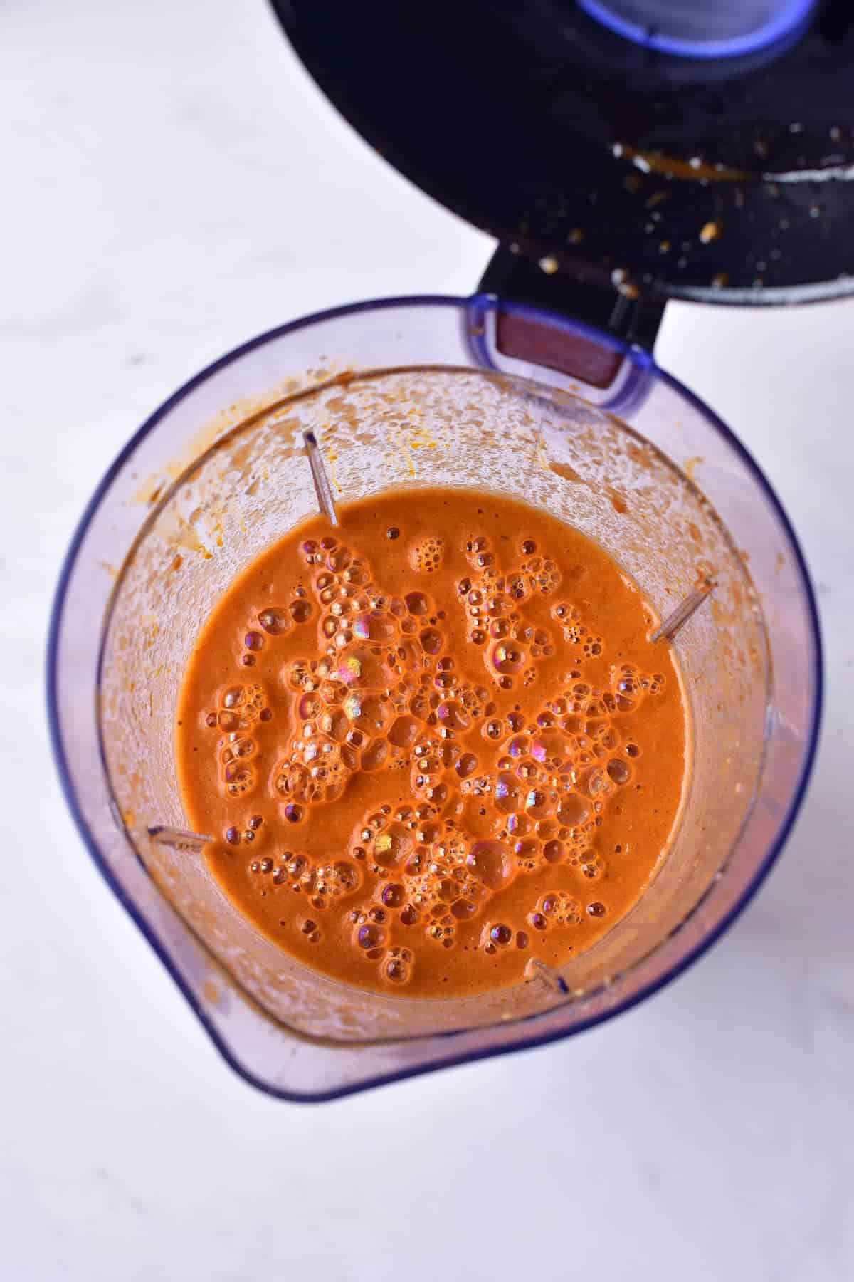 Top-down view of a blender containing a thick, frothy orange-red liquid with bubbles on the surface. The blender lid is partially visible.