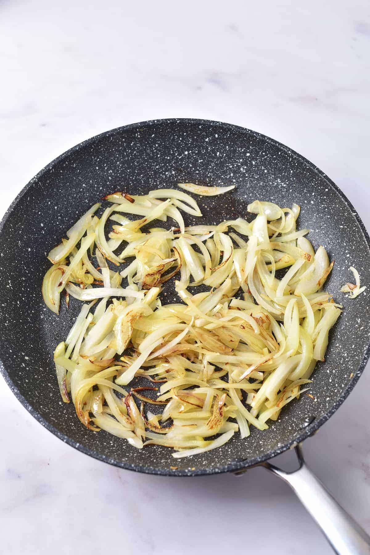 Sliced onions being sautéed and lightly browned in a nonstick frying pan on a white surface.