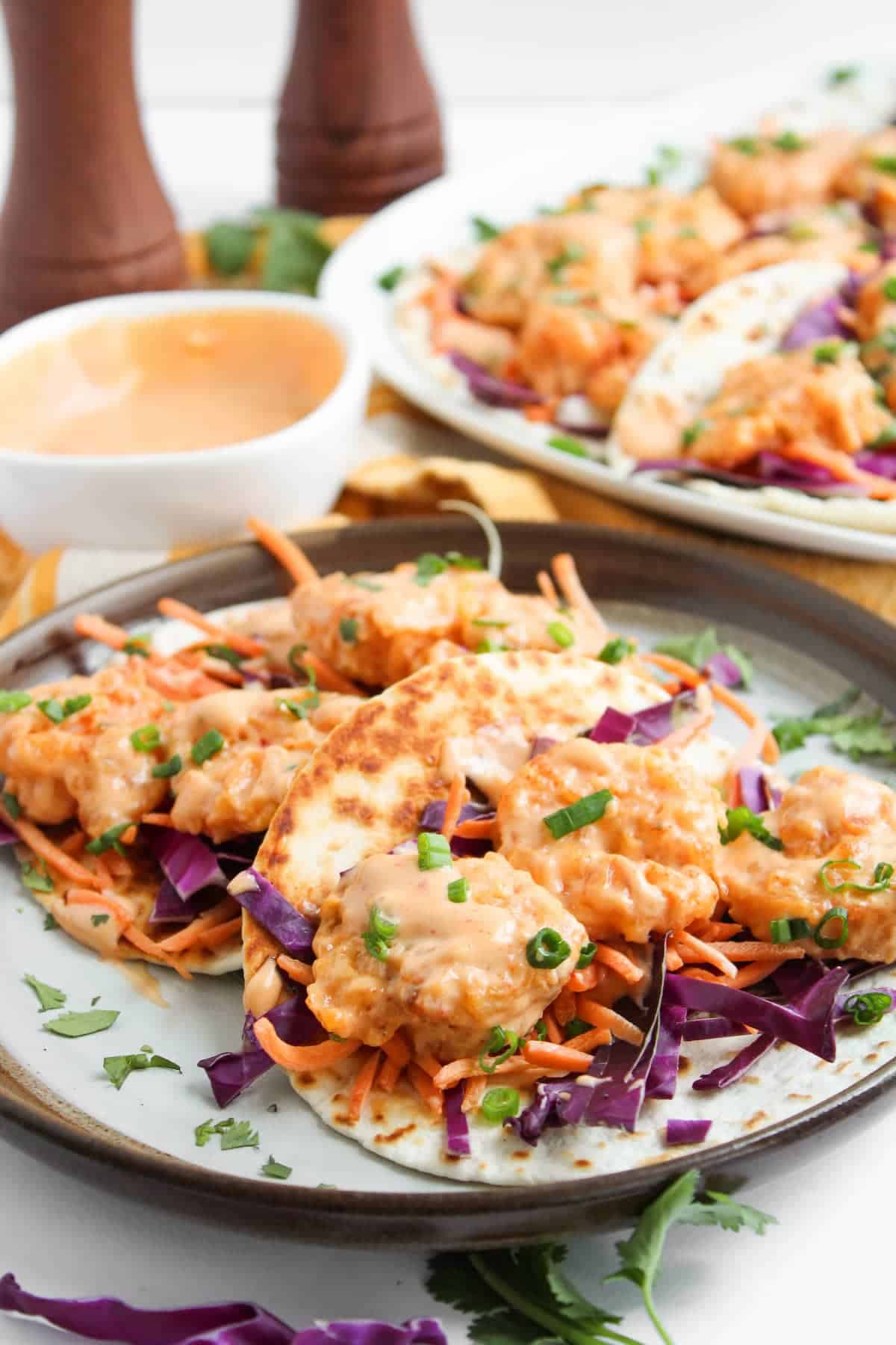 A plate of shrimp tacos with shredded carrots, red cabbage, and chopped green onions, served with a creamy dipping sauce on the side.
