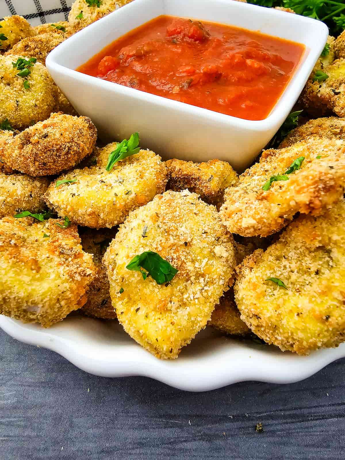 A plate of breaded, baked zucchini slices garnished with herbs, served with a bowl of red marinara dipping sauce.
