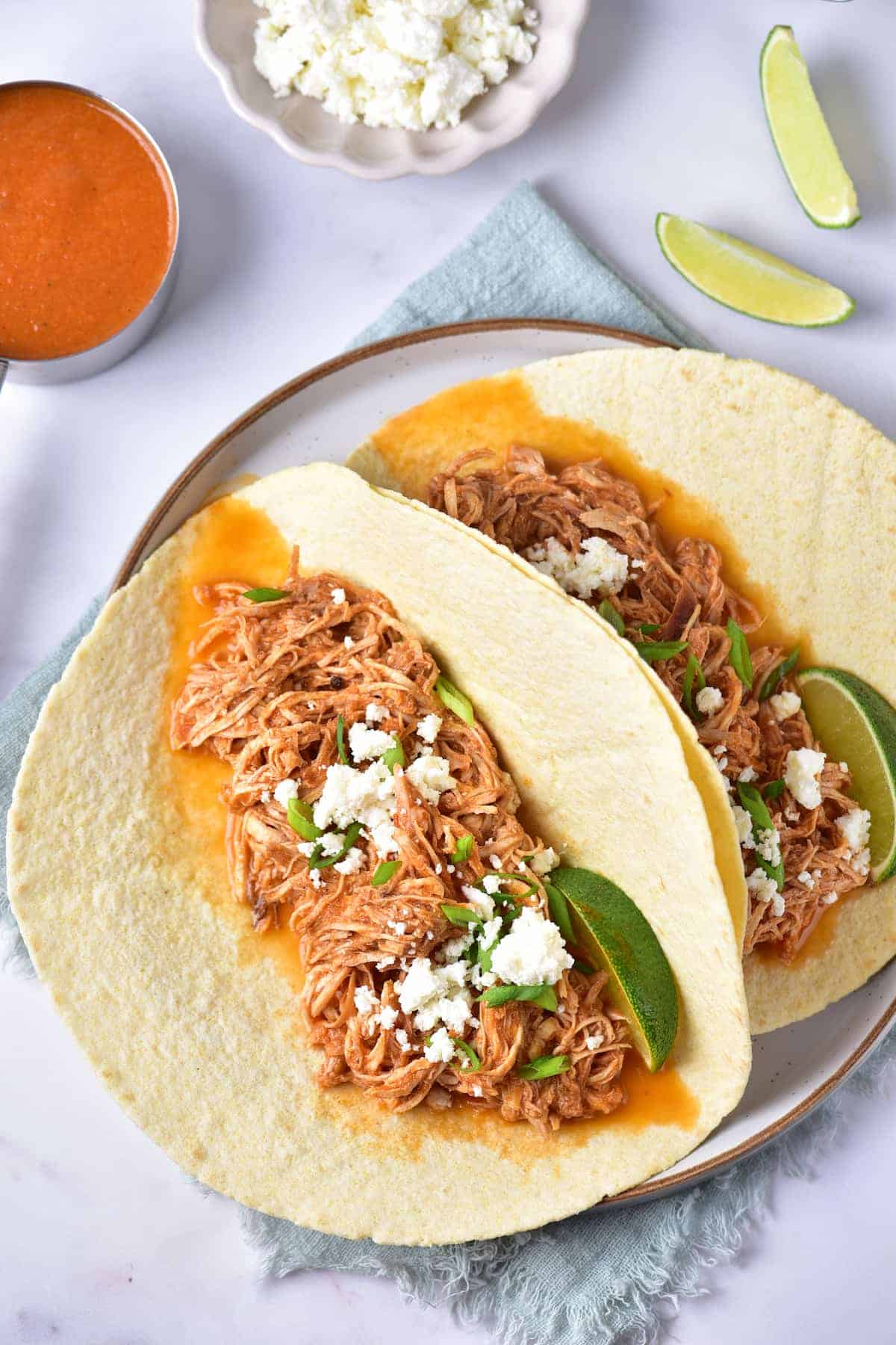 Two flour tortillas filled with shredded chicken, crumbled cheese, green onion, and lime wedges, served on a plate with a side of sauce and extra cheese in the background.