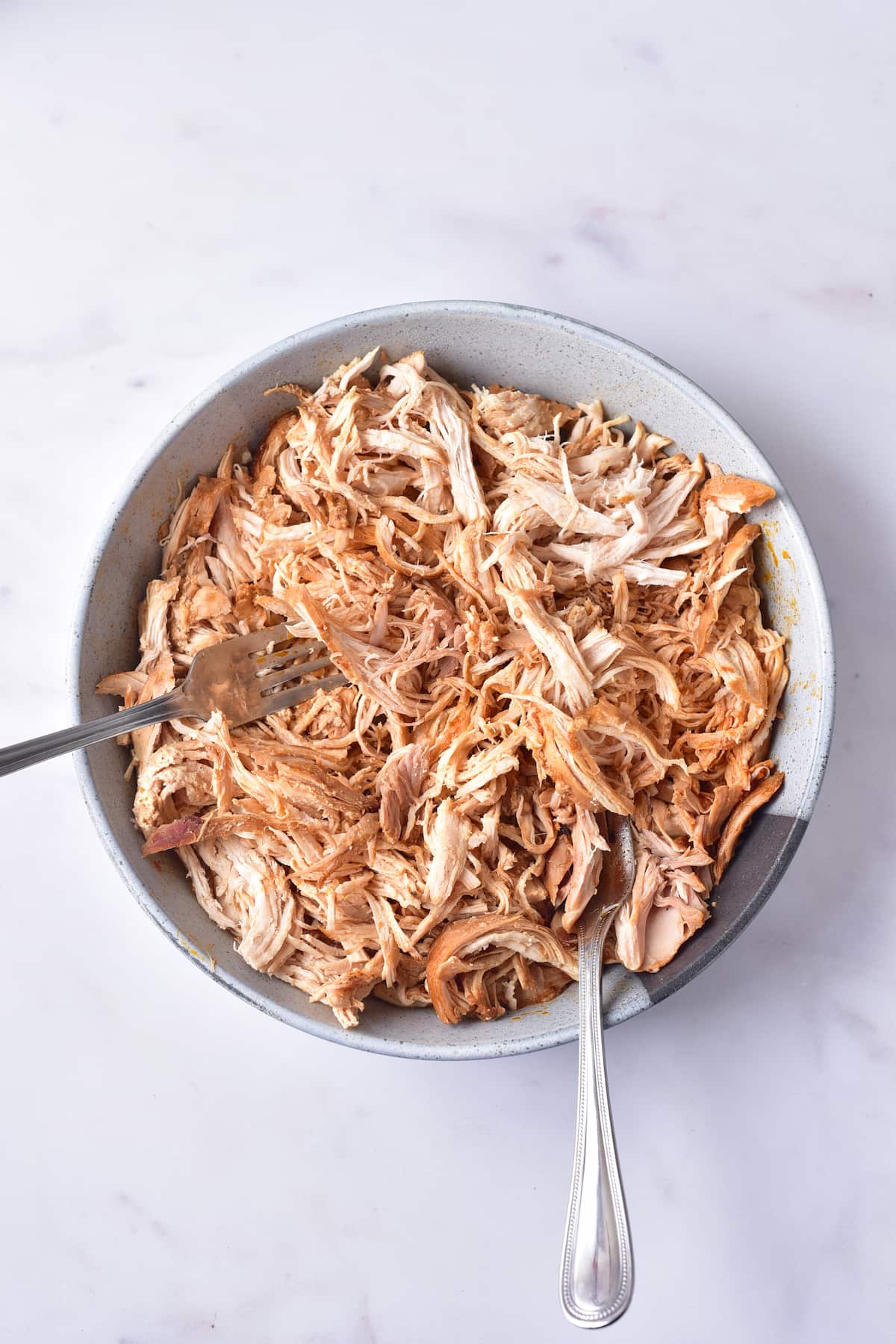 A bowl of shredded cooked chicken with a fork and spoon on a white surface.