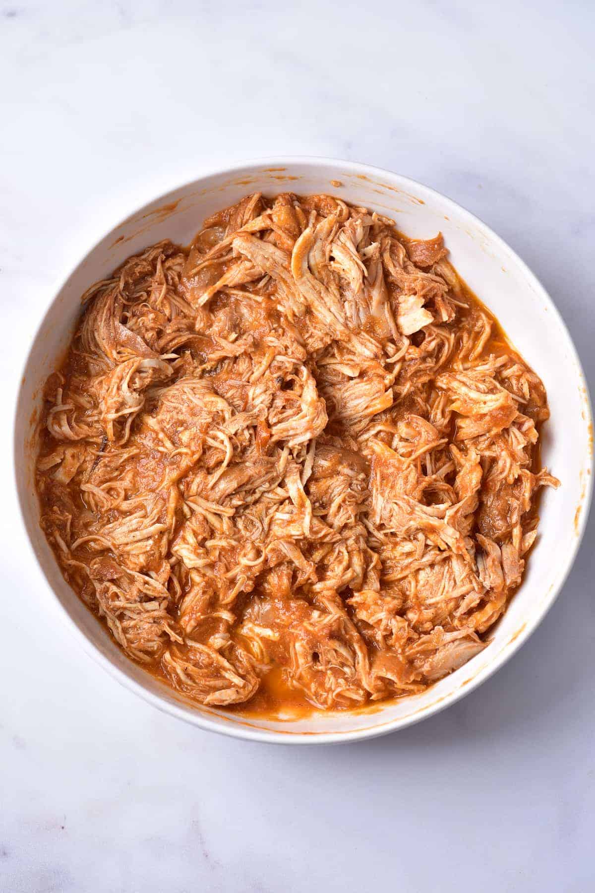 A white bowl filled with shredded chicken coated in a reddish sauce, placed on a white marble surface.