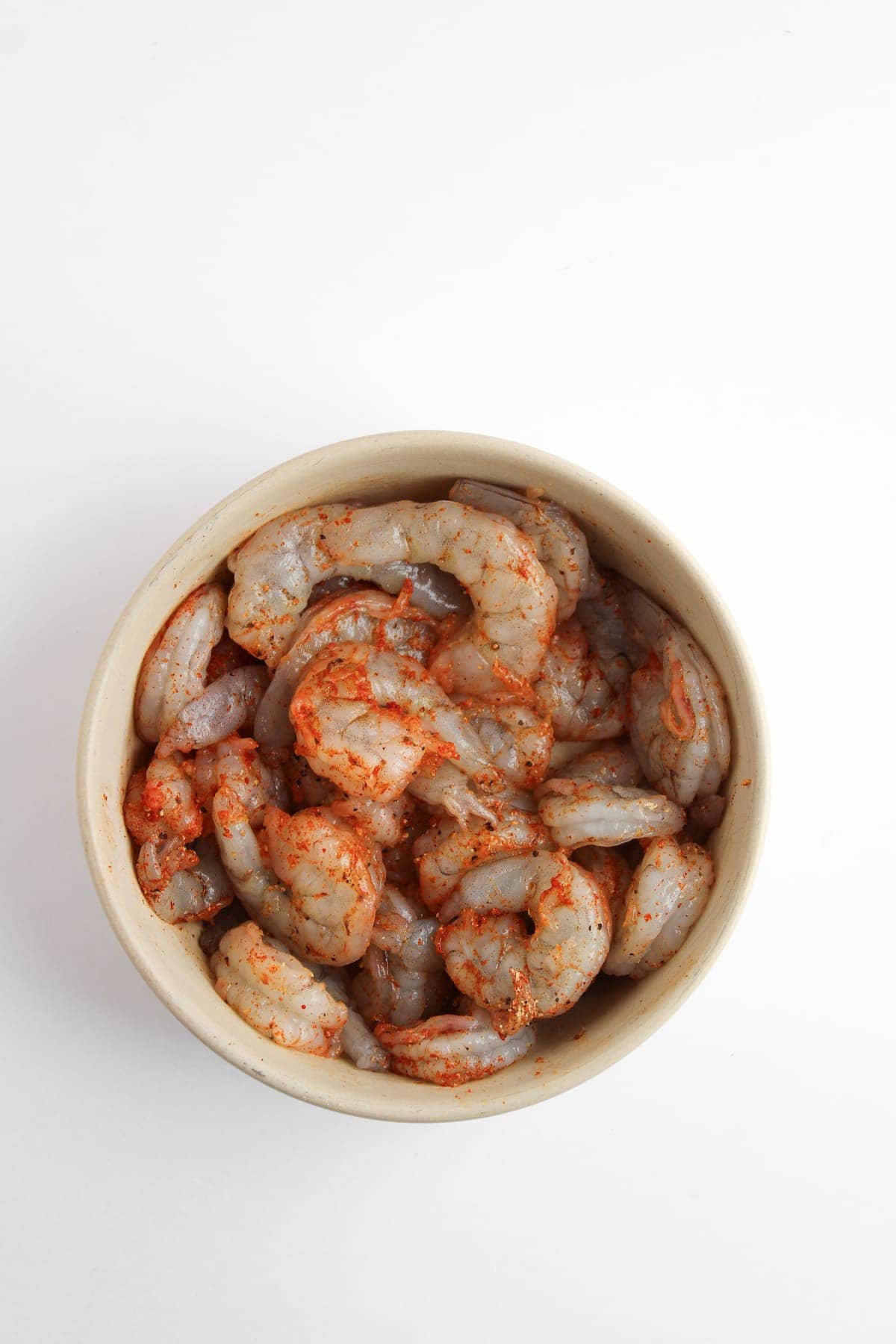 A bowl of raw shrimp seasoned with spices, viewed from above on a white background.