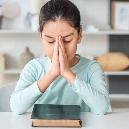 A young girl sits at a table with her hands pressed together in prayer, eyes closed, and a closed book in front of her.