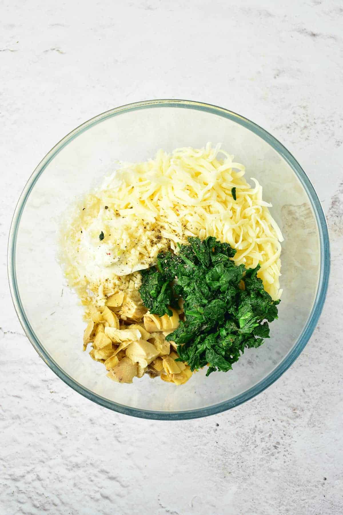 A glass bowl containing shredded cheese, chopped spinach, sliced garlic, and ricotta cheese on a light surface.