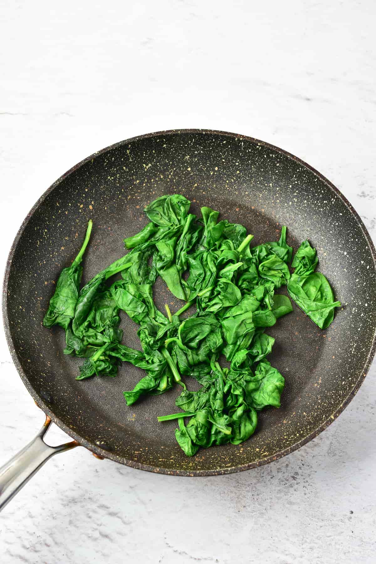 A nonstick frying pan containing cooked spinach leaves on a light-colored surface.