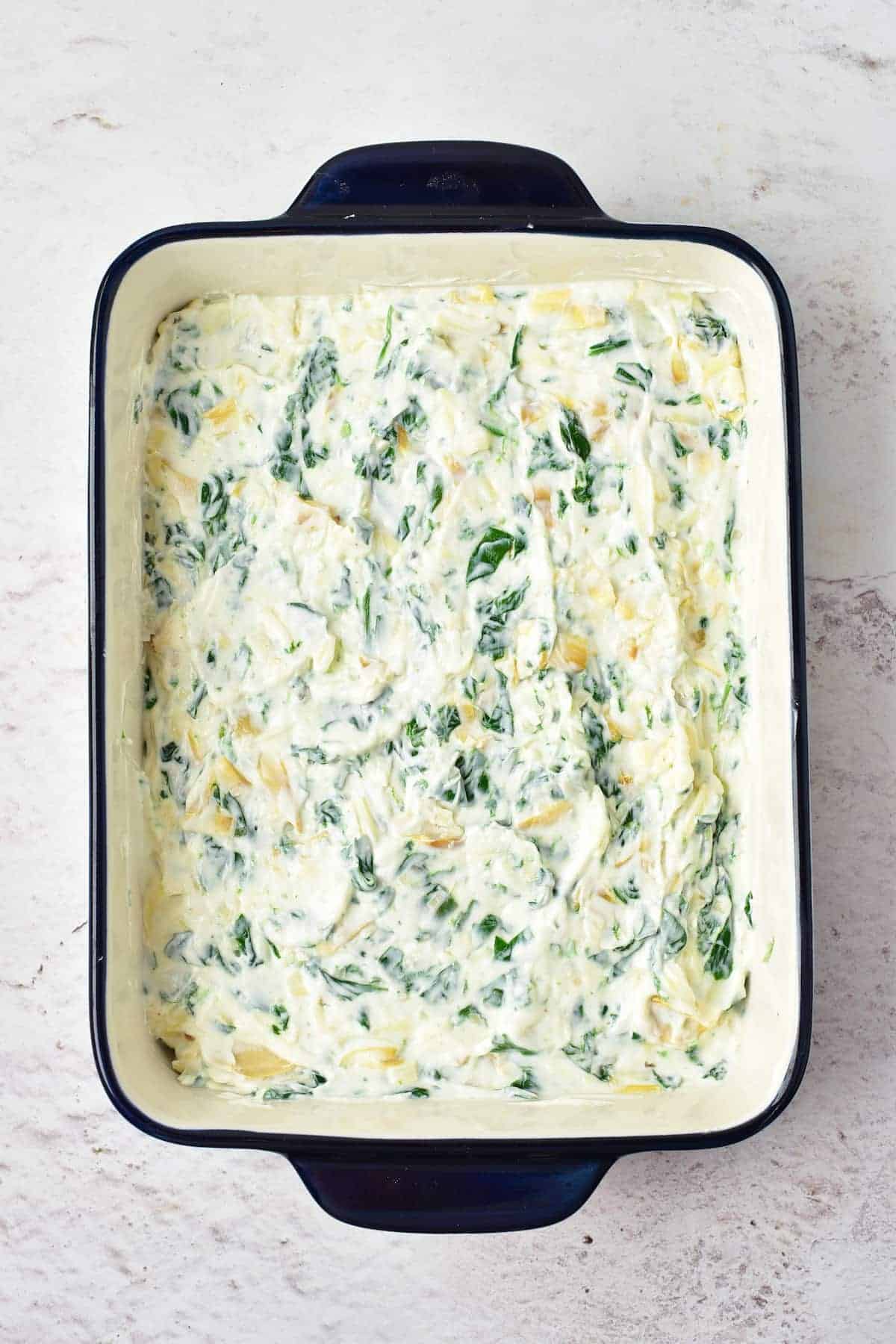 Rectangular baking dish filled with an unbaked mixture of creamy spinach and artichokes on a light-colored surface.