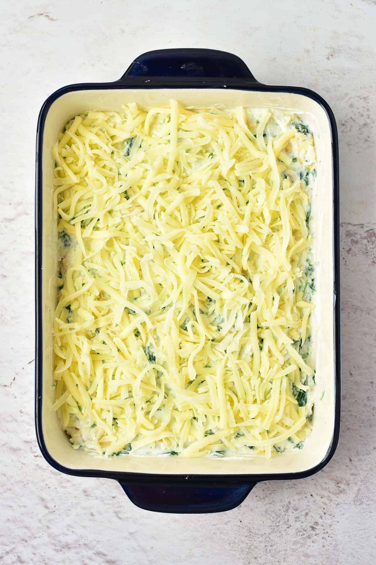 Rectangular baking dish filled with a creamy spinach mixture topped with a layer of shredded cheese, ready to be baked.