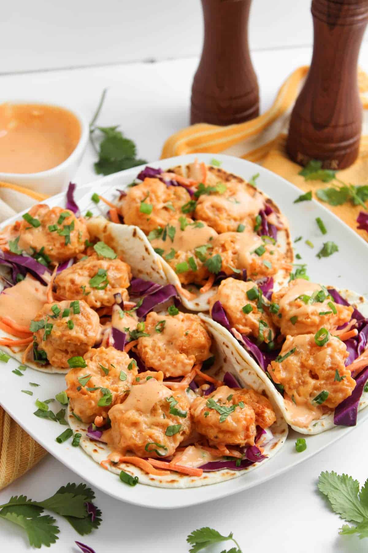 A plate of tacos filled with shrimp, shredded cabbage, sauce, and chopped herbs, with a bowl of sauce, cilantro leaves, and two pepper mills in the background.