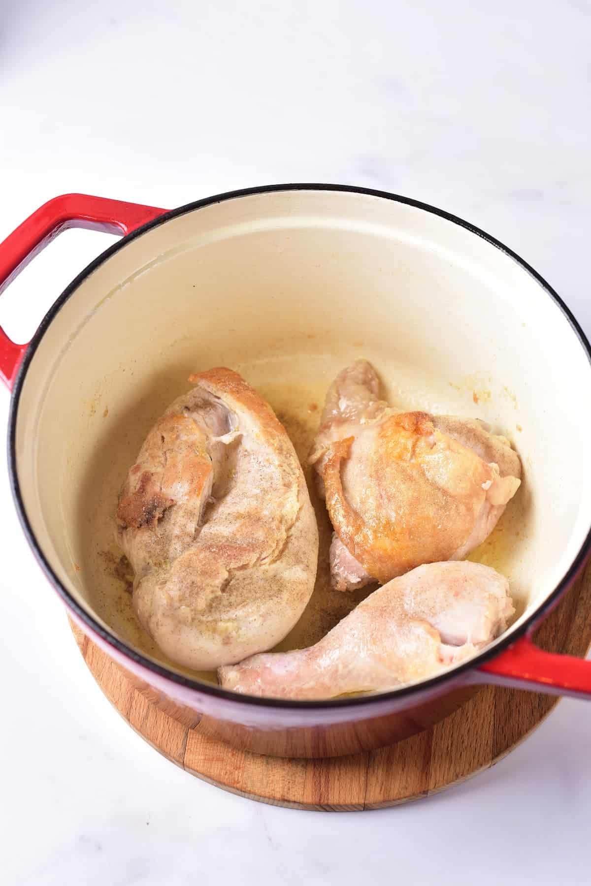 Three pieces of browned chicken cooking in a red pot on a wooden trivet, set on a white surface.