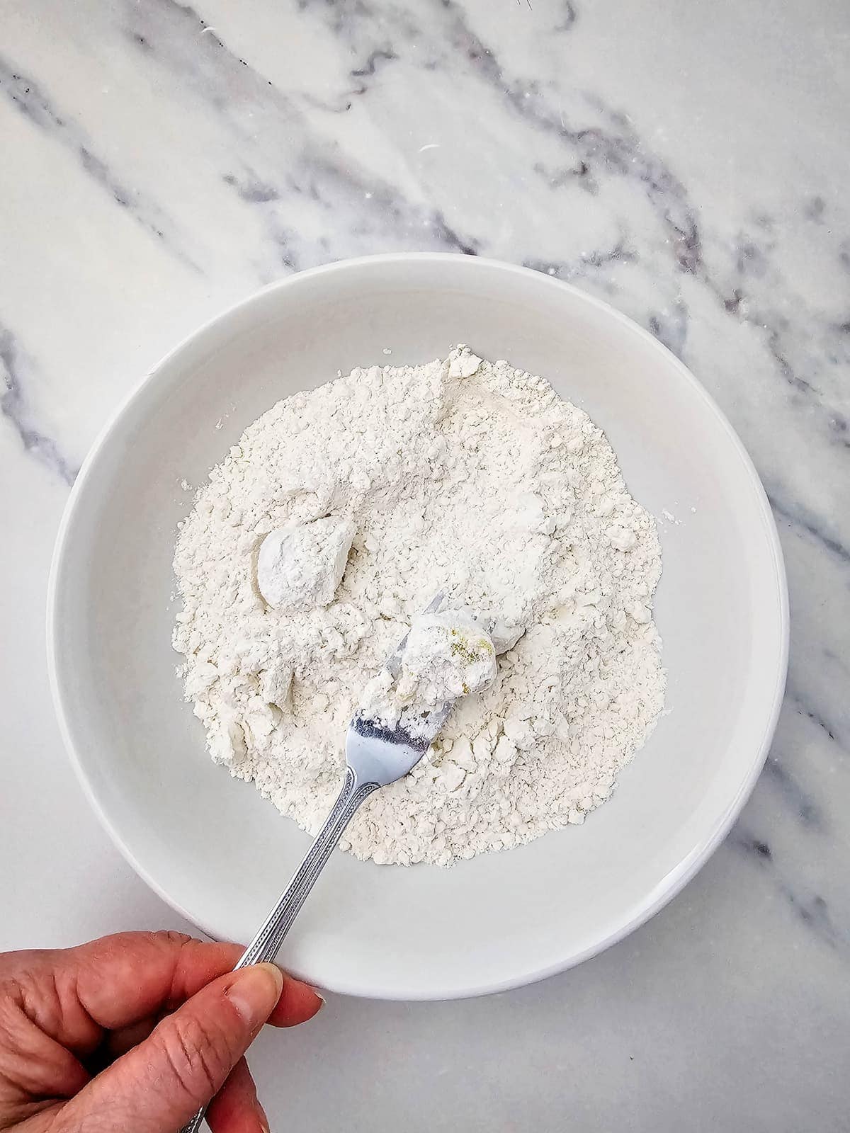 A hand holding a fork mixes white flour in a white bowl placed on a marble countertop.