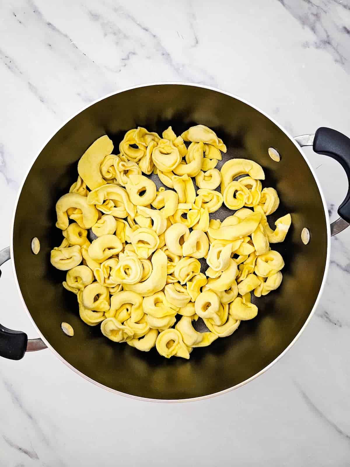 A black pot filled with uncooked tortellini pasta sits on a white marble countertop.
