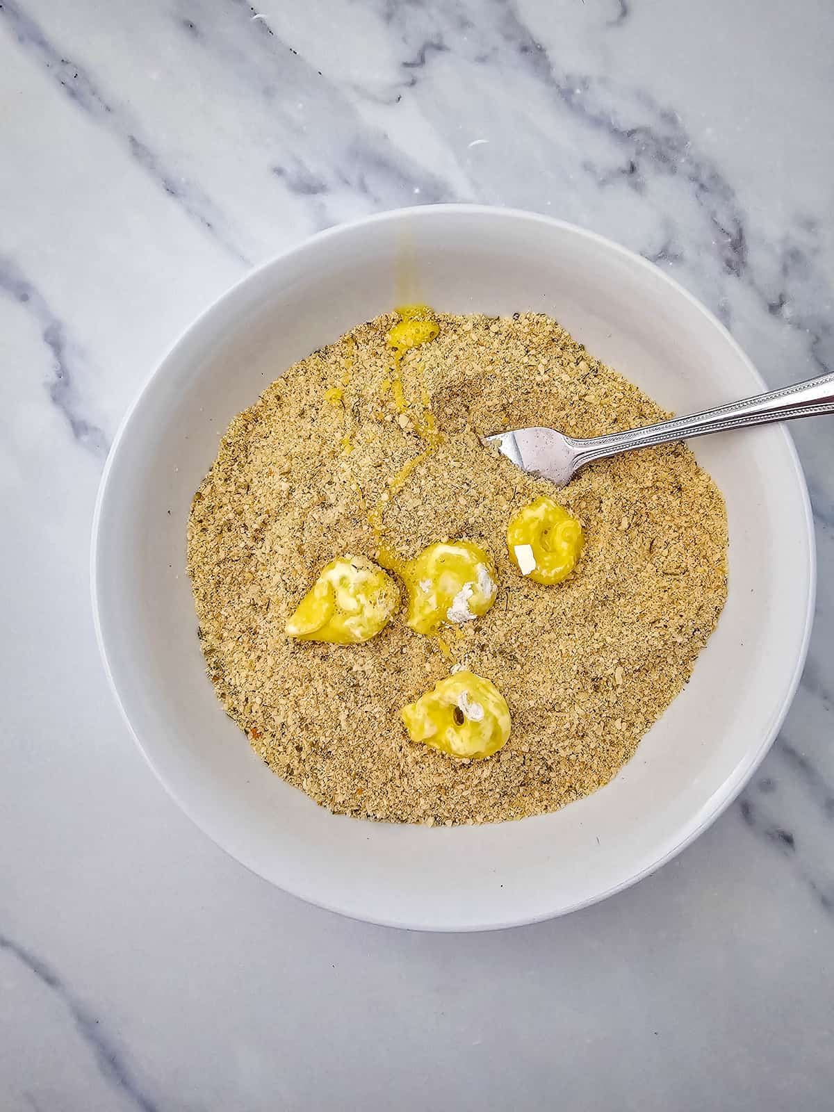 A white bowl containing dry breadcrumb mixture with dollops of beaten egg and a fork, placed on a marble countertop.