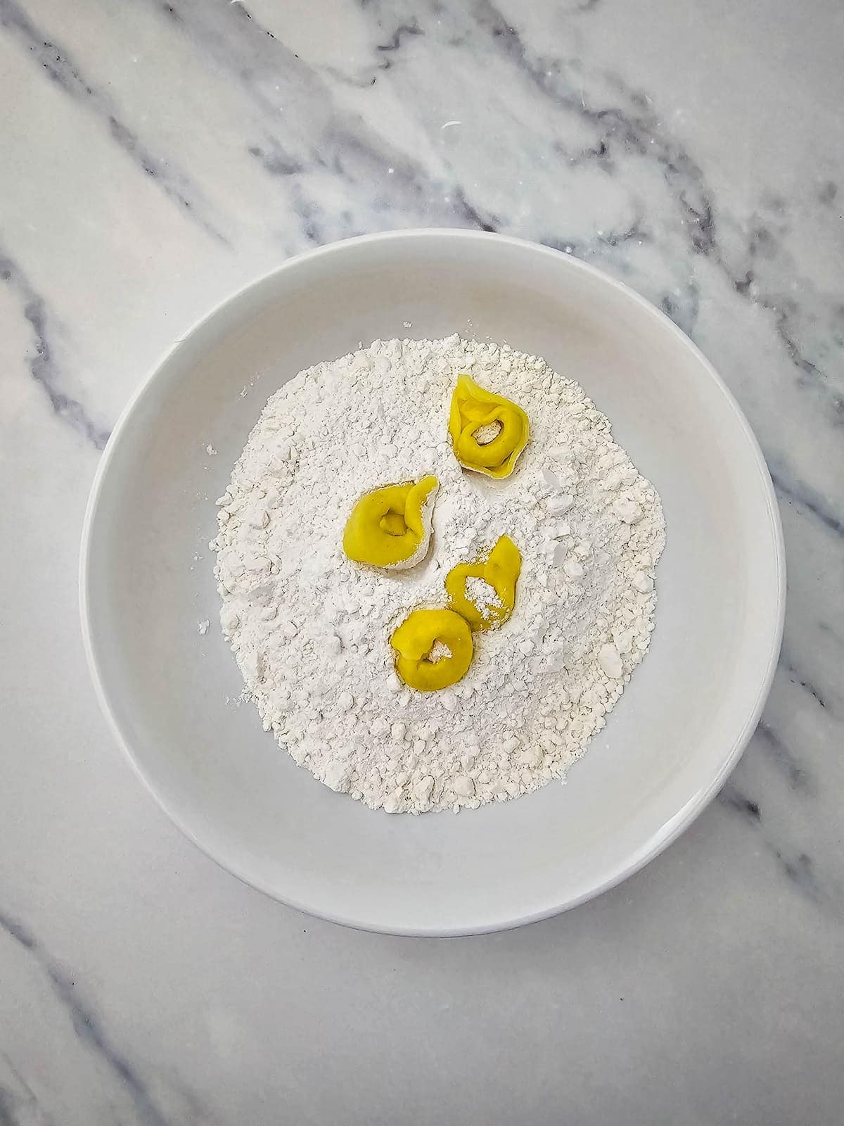 A white bowl with flour and four pieces of uncooked tortellini on a light marble surface.