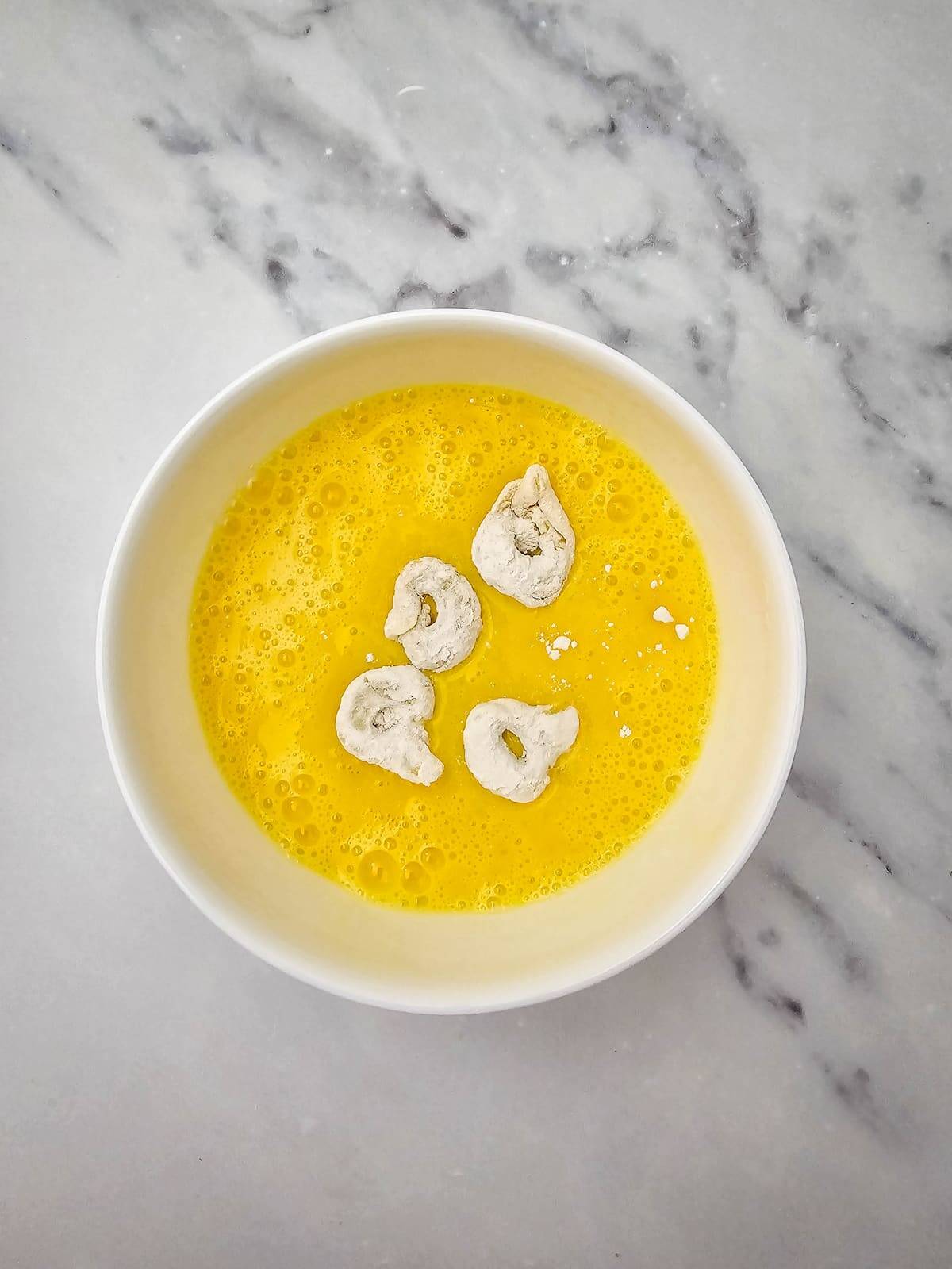 A white bowl containing beaten eggs with four pieces of flour-coated food on a marble surface.
