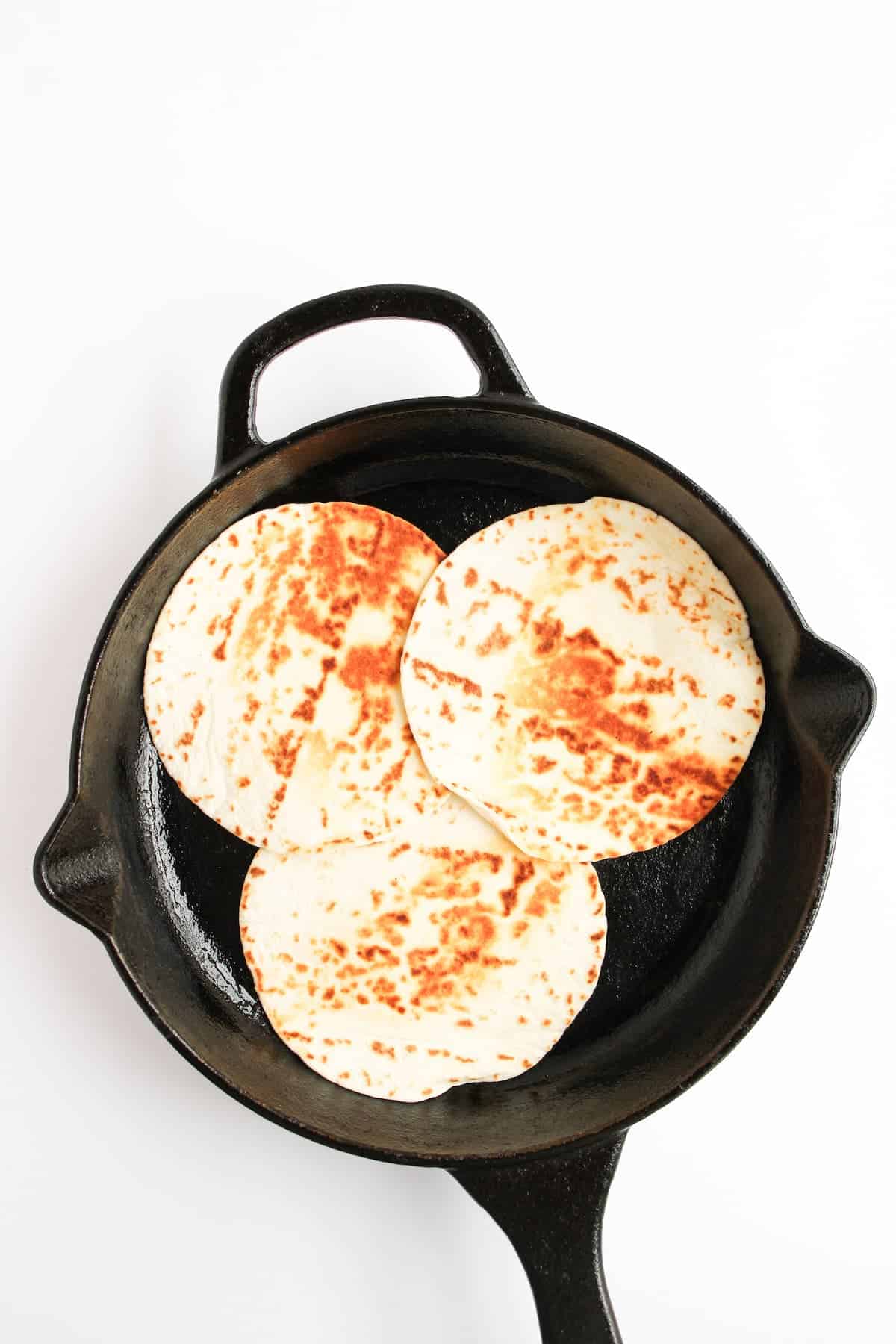 Three tortillas are heating in a black cast iron skillet on a white background.