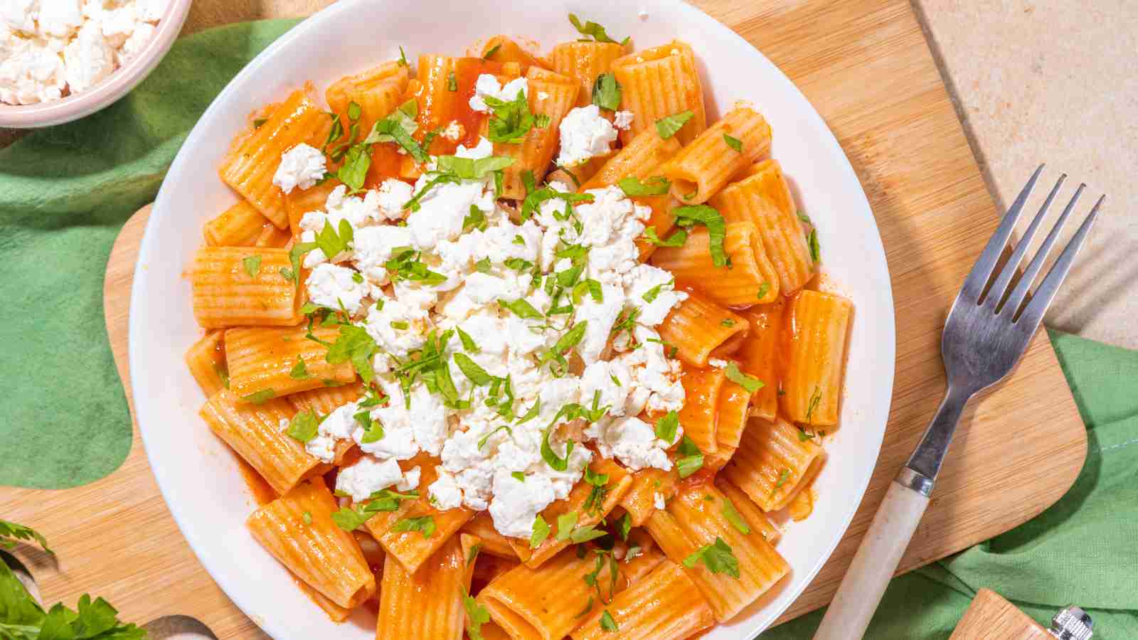 A plate of rigatoni pasta with tomato sauce, topped with crumbled white cheese and chopped fresh herbs, placed on a wooden board with a fork beside it.