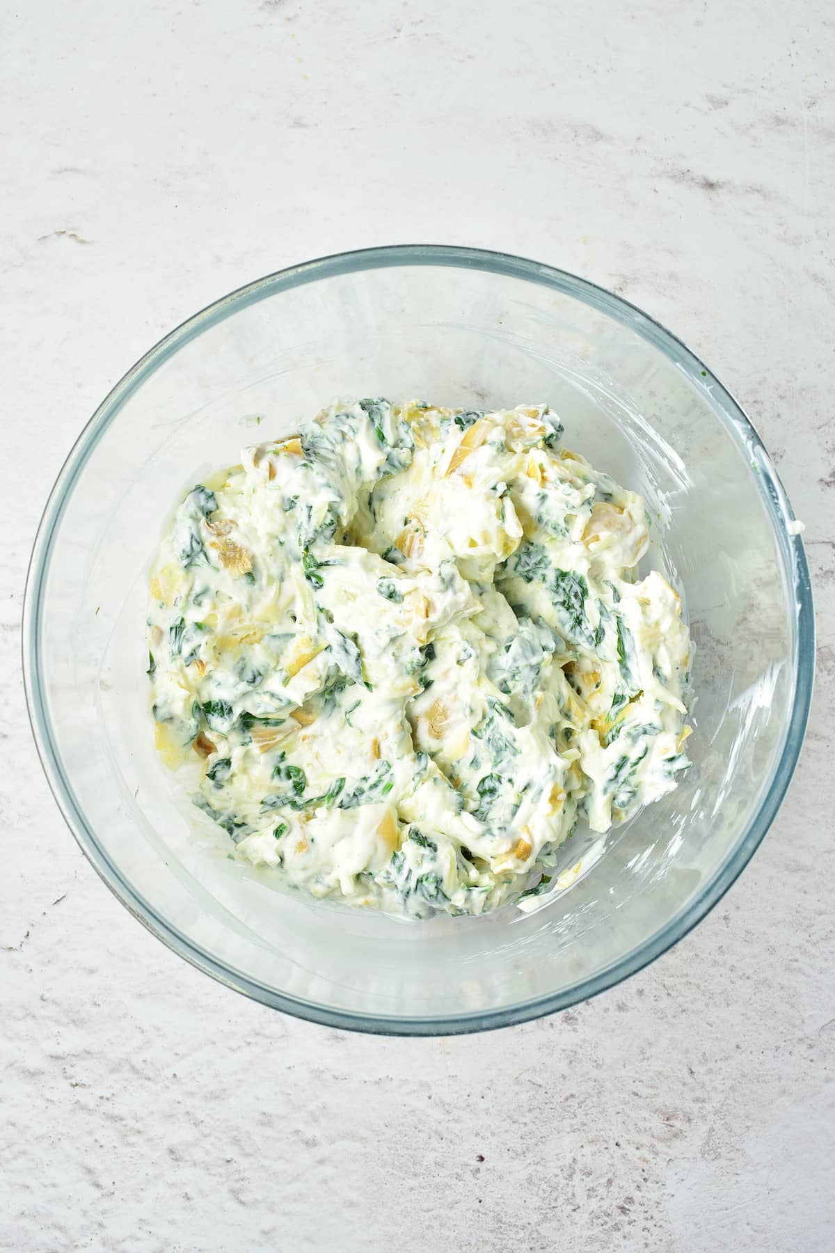 A glass bowl filled with creamy spinach and artichoke dip sits on a white textured surface.