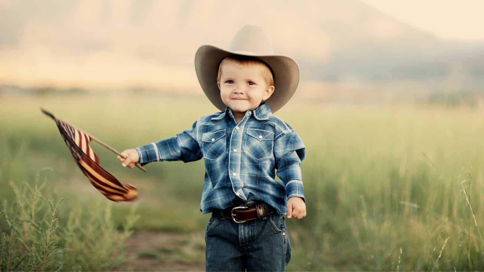 A young boy in a plaid shirt and cowboy hat stands in a grassy field, holding a small American flag.