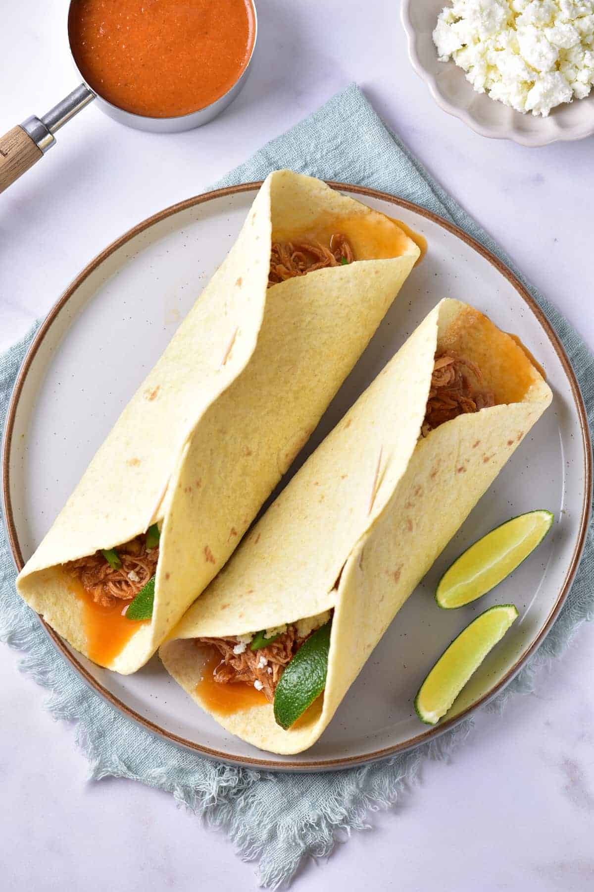 Two tortilla wraps filled with shredded meat and vegetables on a plate, served with lime wedges. A bowl of sauce and a dish of crumbled cheese are nearby.