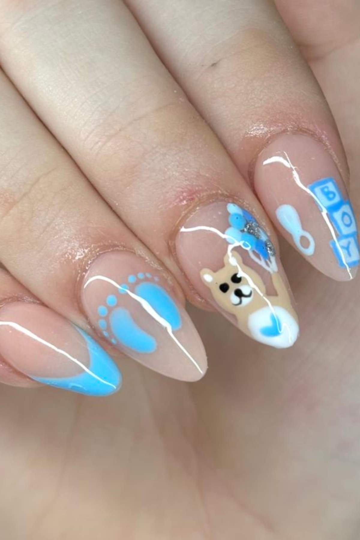 A hand with almond-shaped nails featuring baby boy-themed nail art, including blue footprints, a teddy bear, a rattle, blocks spelling "BOY," and blue French tips.