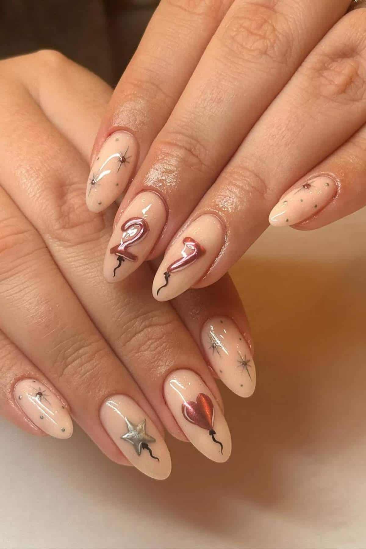 A hand with almond-shaped nails painted nude, featuring balloon designs with the numbers "2" and "7," a heart, a star, and small silver star accents.