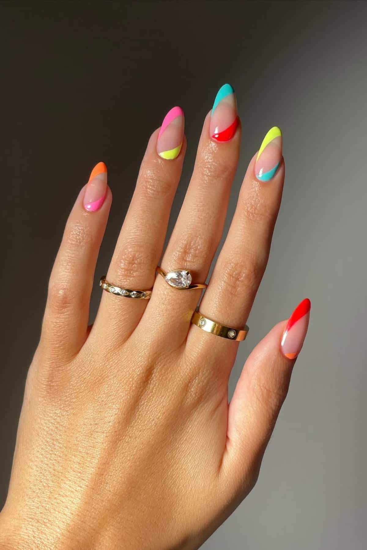 A hand with almond-shaped nails painted in a colorful, diagonal French tip design, wearing four gold rings.
